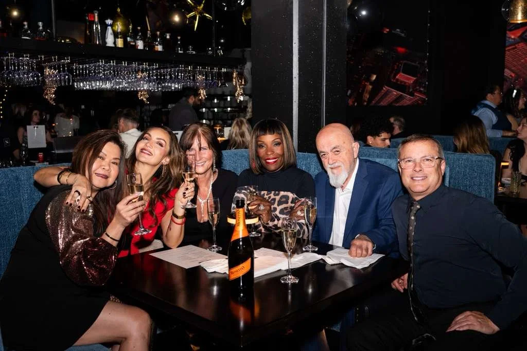 Group of six people celebrating at a restaurant table with champagne glasses and a bottle, smiling and enjoying the night.