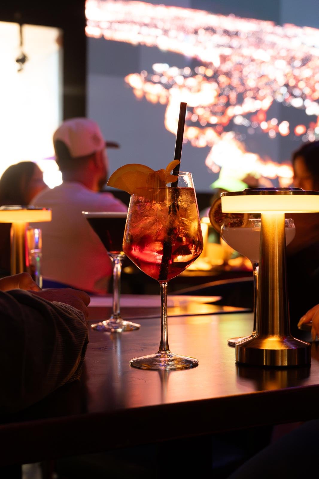 A colorful drink garnished with a lemon slice and a straw on a bar table, with other drinks and people in the background at a dimly lit lounge or restaurant with a large screen showing colorful lights or a cityscape.
