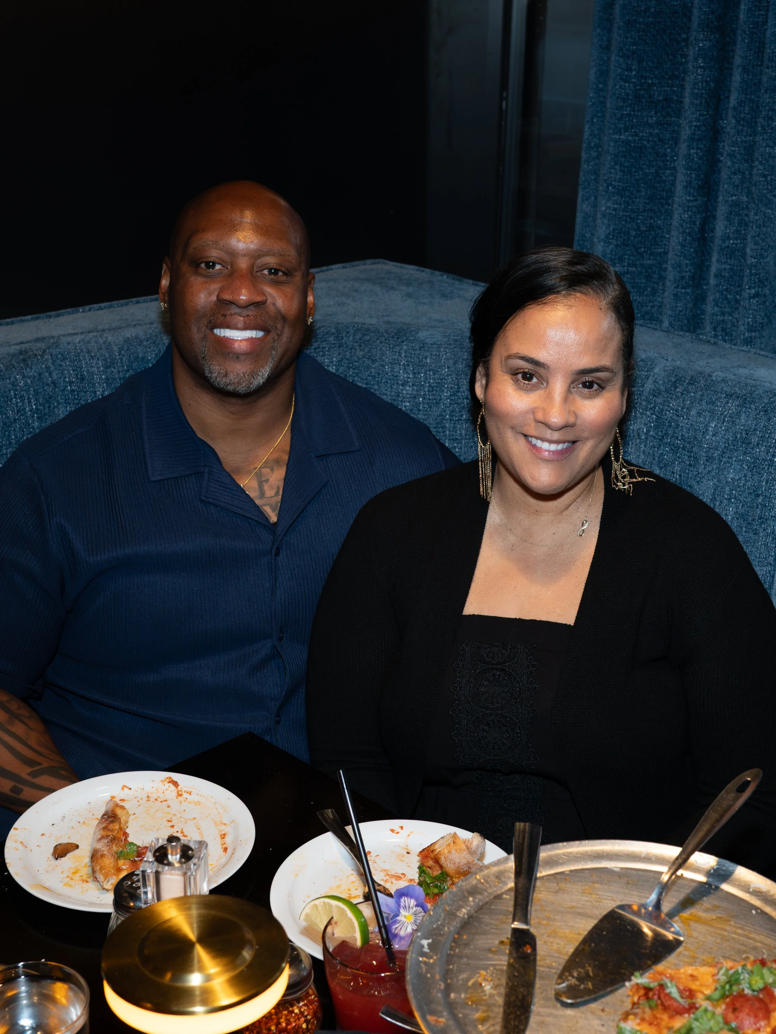 A man and a woman sitting at a table in a restaurant or dining setting, smiling at the camera. The table has plates with partially eaten pizza and a drink with a lime wedge, with some food and utensils visible around.