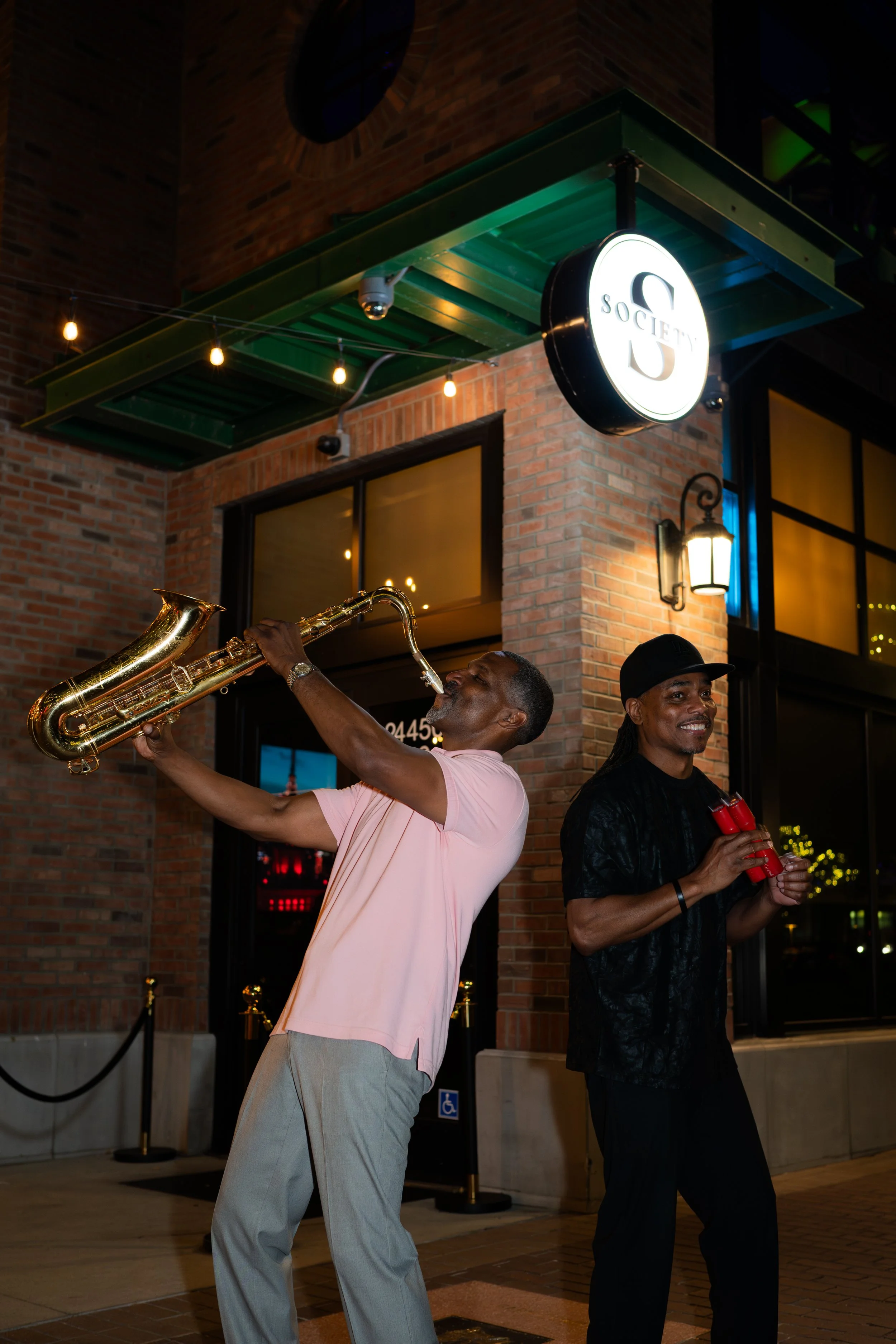 Two musicians performing outside a brick building at night, one playing the saxophone and the other holding a drink.