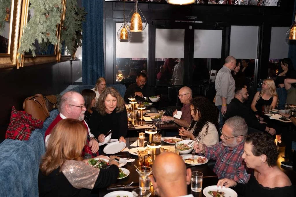 People dining at a restaurant table with plates of food, illuminated by warm hanging lights, and a window showing reflections of more diners.