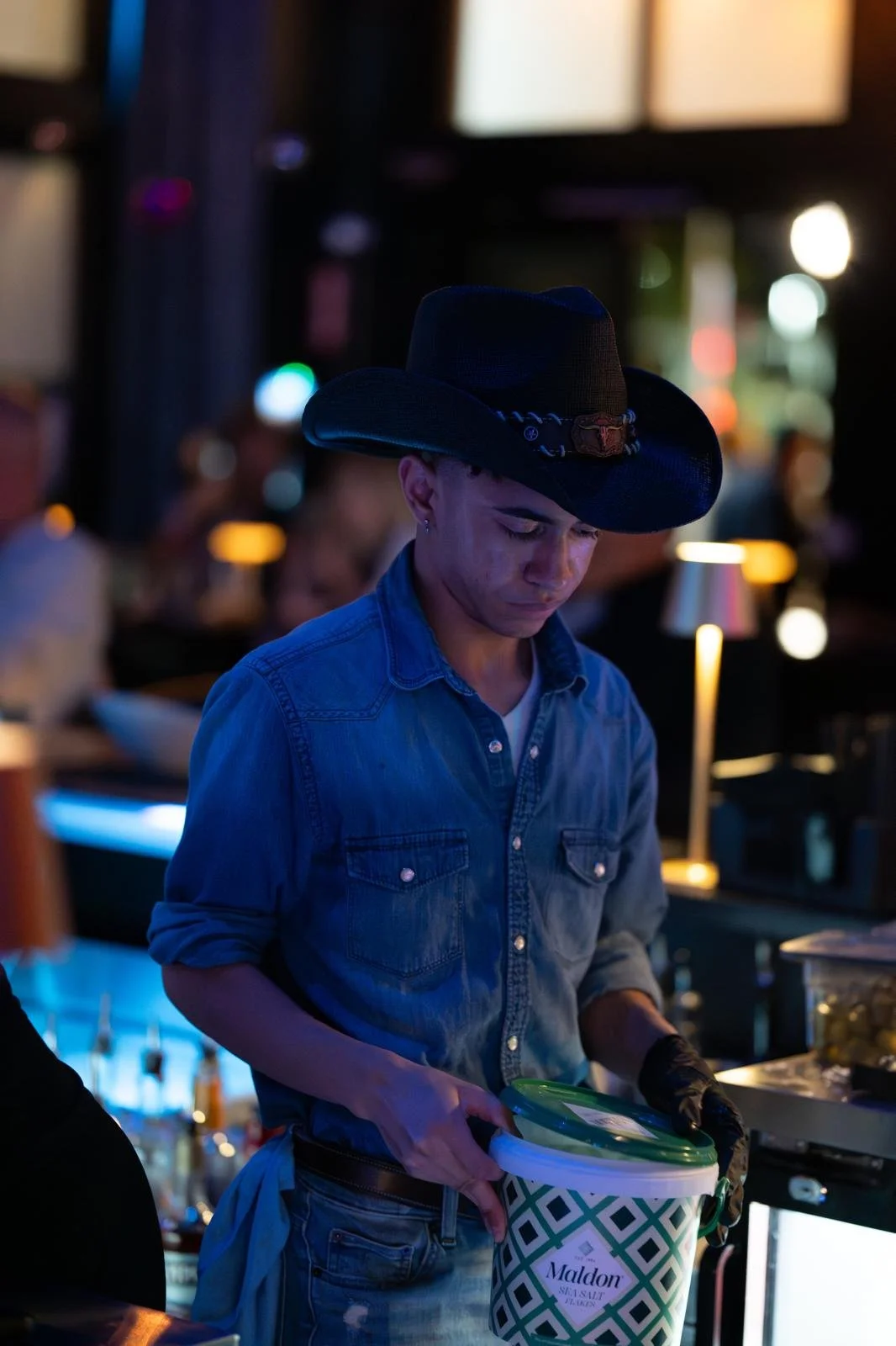 A young man wearing a black cowboy hat, denim shirt, and black gloves, holding a green and white container labeled 'Maldon' in a dimly lit restaurant or bar.