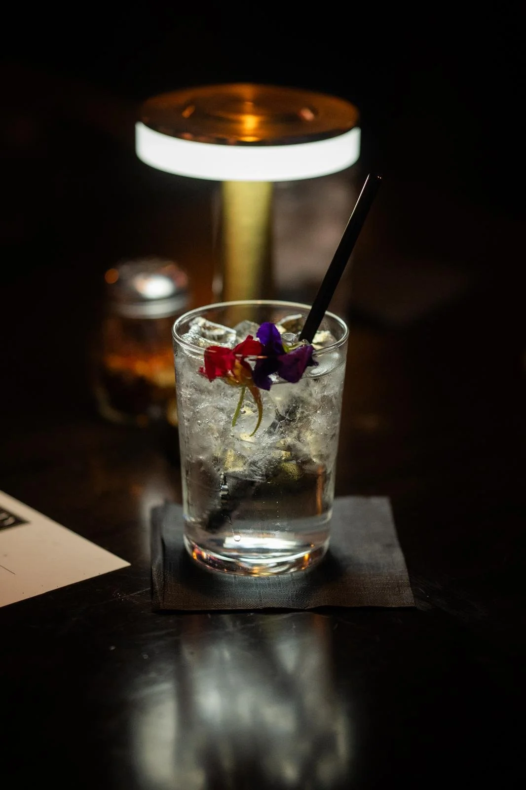 A glass of iced water with a black straw and purple and red flowers garnishing it, placed on a black napkin on a dark table with a warm, dim light above.