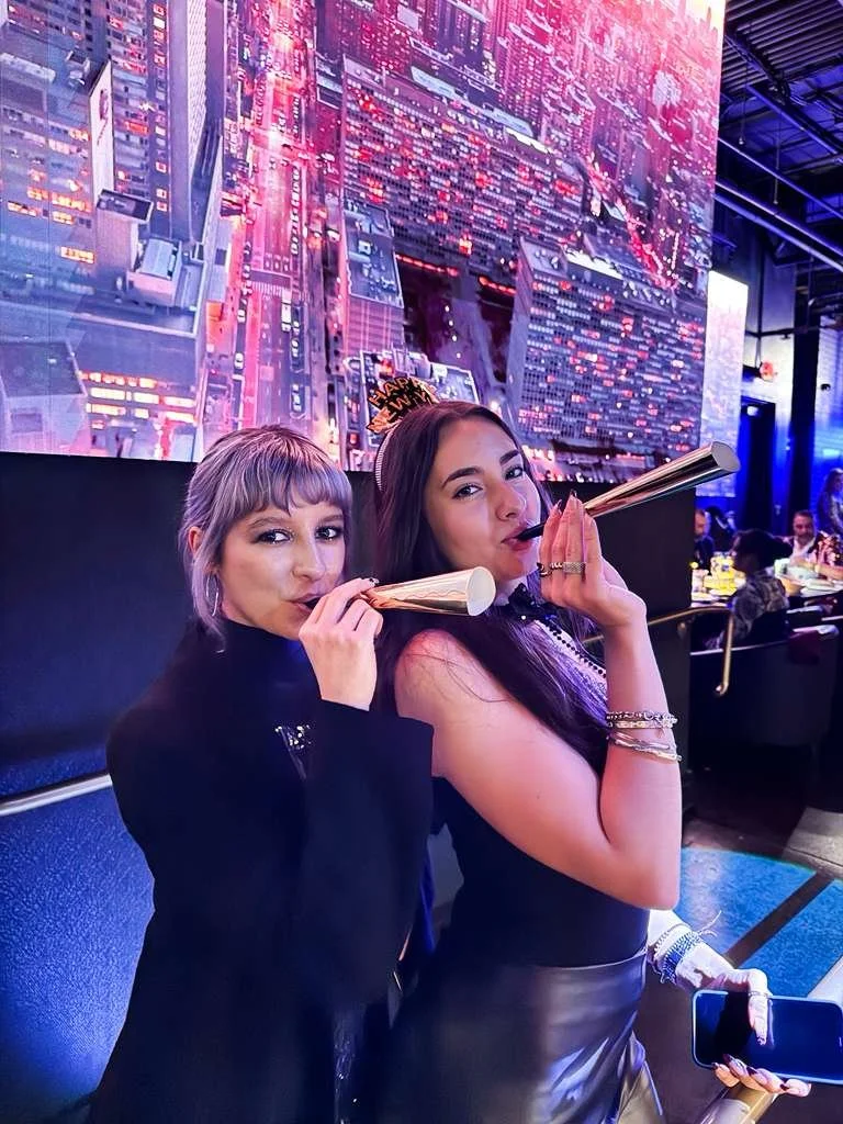 Two women celebrating a New Year's Eve party, wearing party hats, blowing party horns, and posing in front of a vibrant cityscape digital display at a nightclub or event venue.