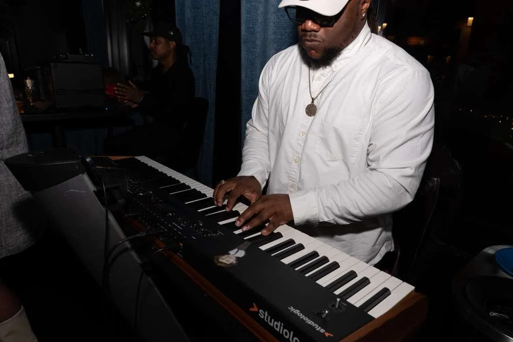 A man wearing sunglasses and a white shirt playing a keyboard in a dimly lit room, with another person in the background sitting at a desk.