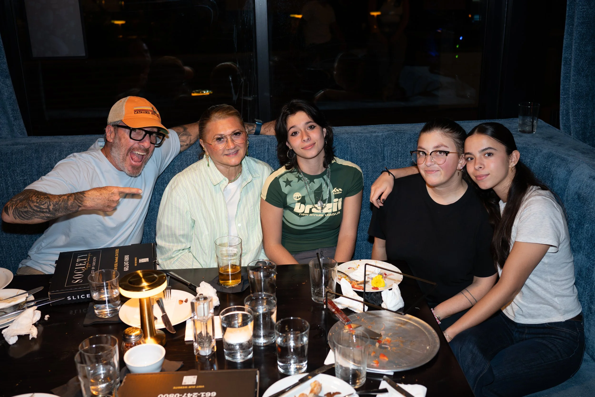 A group of five people sitting at a restaurant table, smiling and posing for a photo. The table has plates, glasses, and used food. The background is dimly lit with reflections on the window.