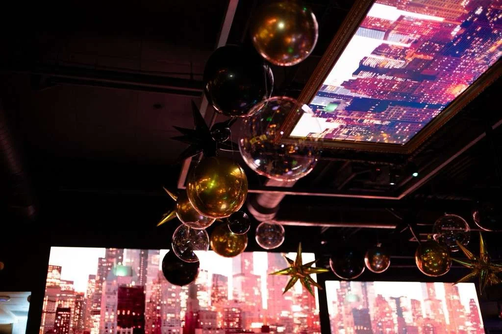 Decorative hanging Christmas ornaments, including gold and black baubles and golden star-shaped decorations, with cityscape and digital city images projected on screens and ceiling in the background.