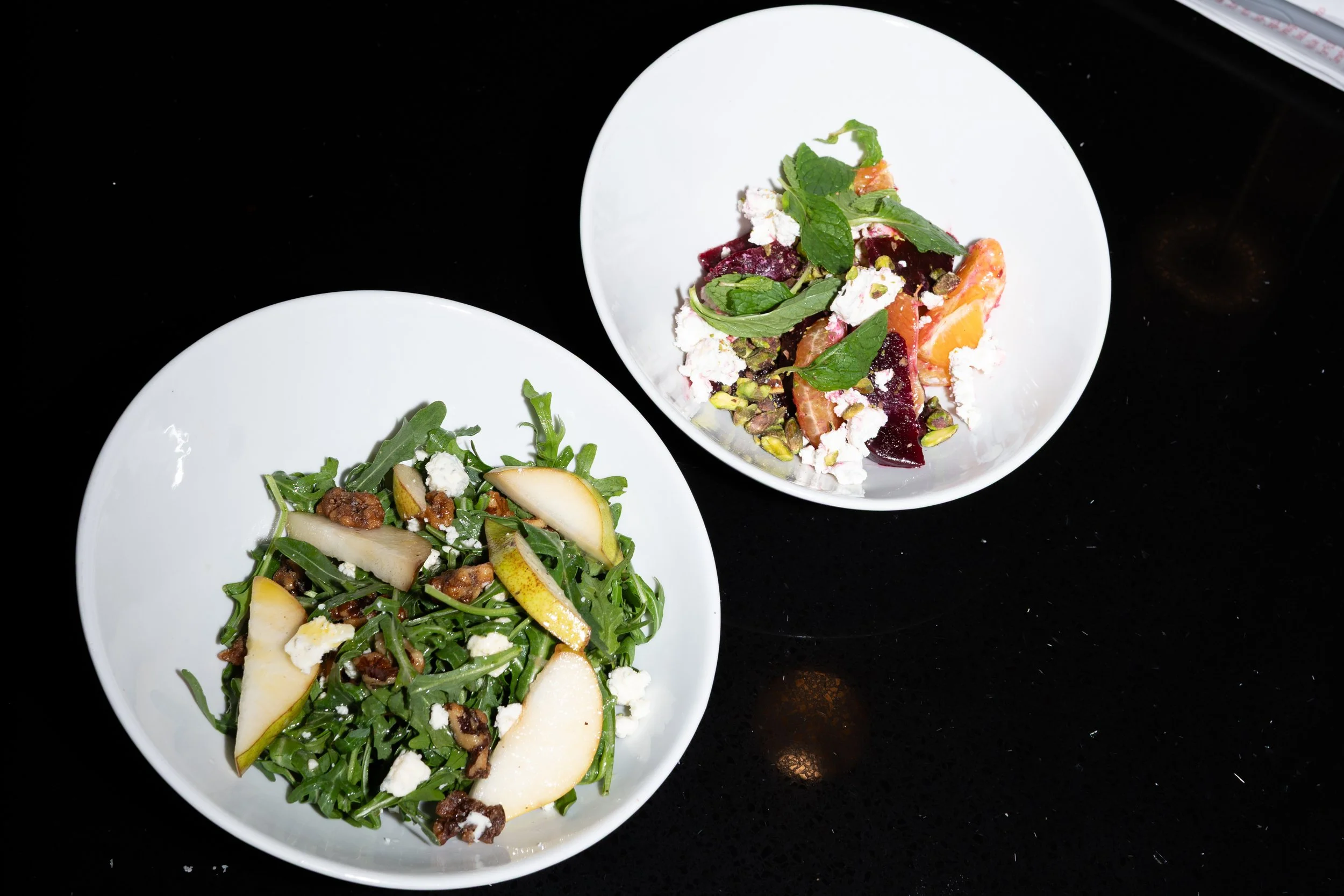 Two white bowls on a black surface, one containing a pear and walnut salad with greens, cheese, and nuts, and the other containing a fruit and herb salad with citrus segments, berries, mint, and crumbled cheese.