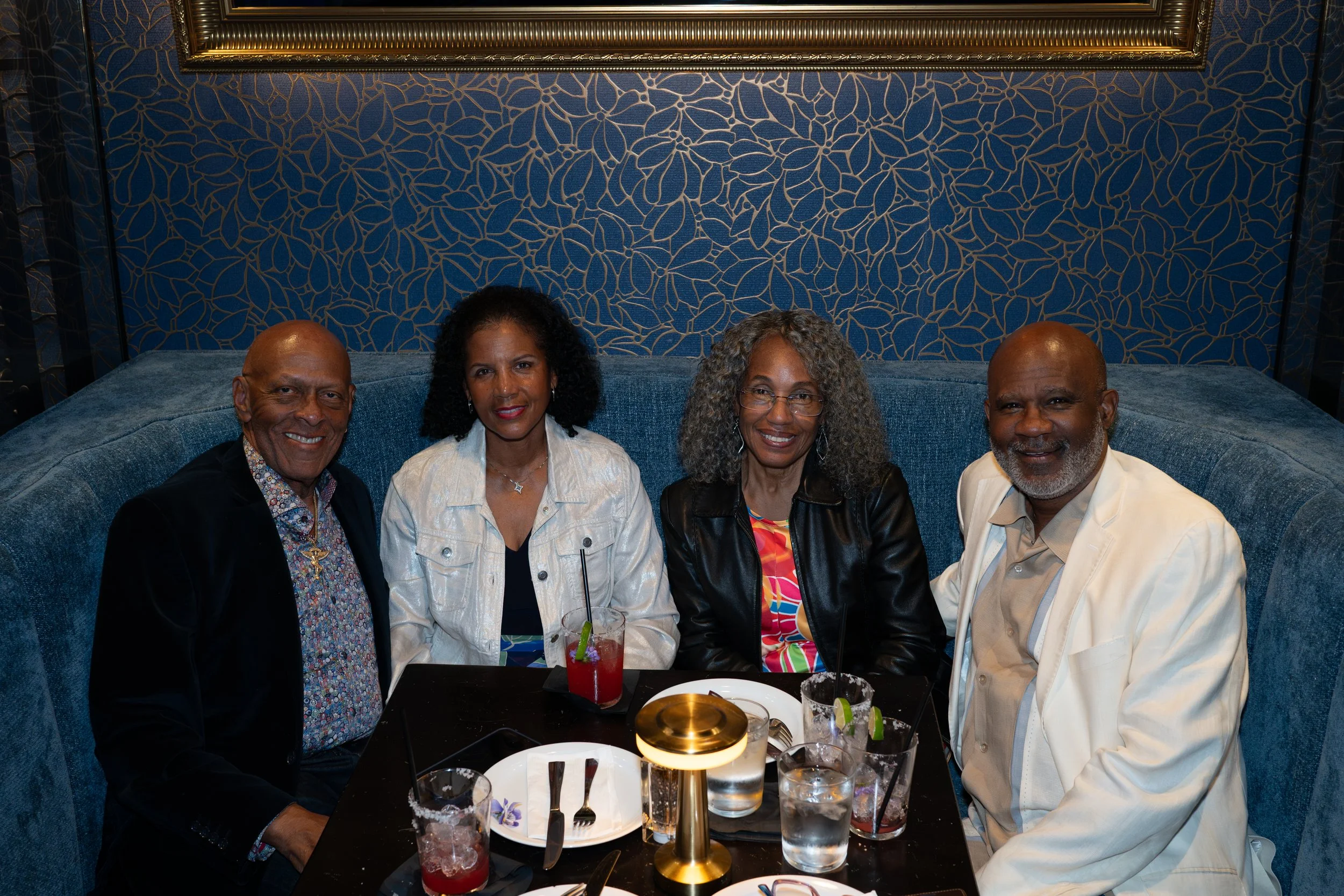Four adults sitting at a restaurant table with drinks and plates of food, in a dimly lit setting with blue and gold decor.