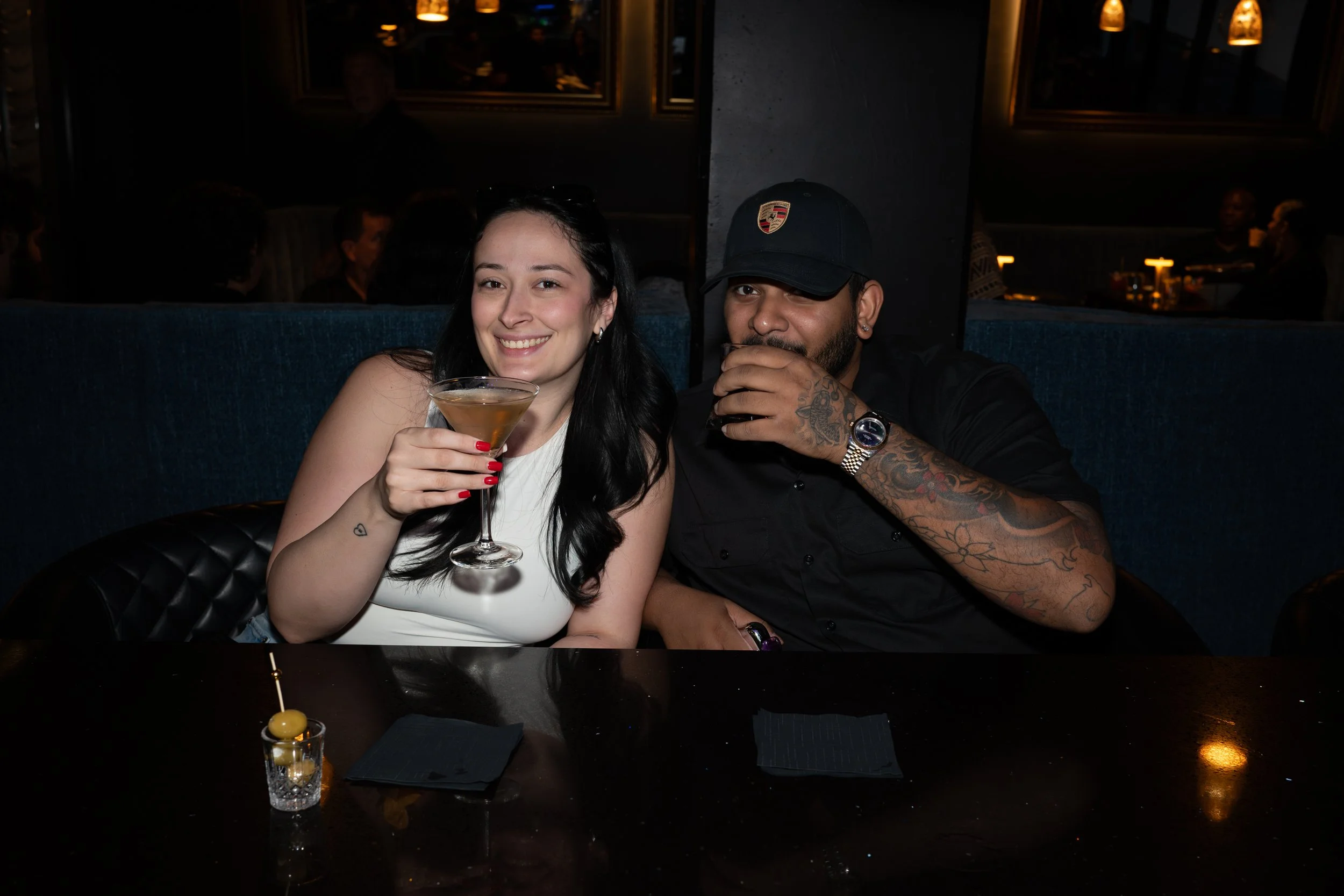 A woman with dark hair, wearing a white top, is smiling and holding a drink in a glass, sitting beside a man with tattoos, a dark baseball cap, and earrings, in a dimly lit restaurant or bar. They are sitting at a table with an appetizer and black na