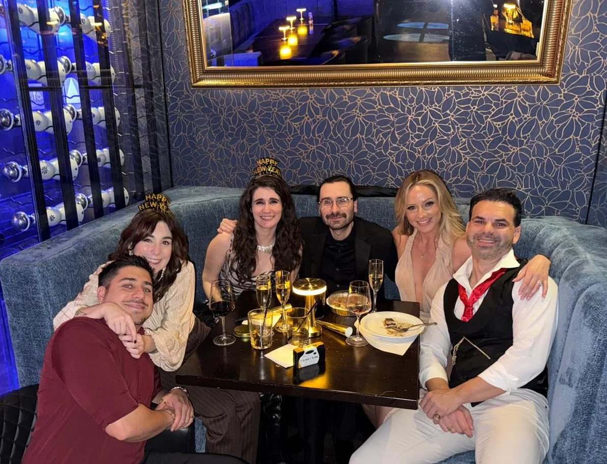 Group of six people celebrating New Year's at a restaurant, wearing 'Happy New Year' headbands, with champagne glasses on the table, in a cozy booth with patterned wallpaper and a large mirror.