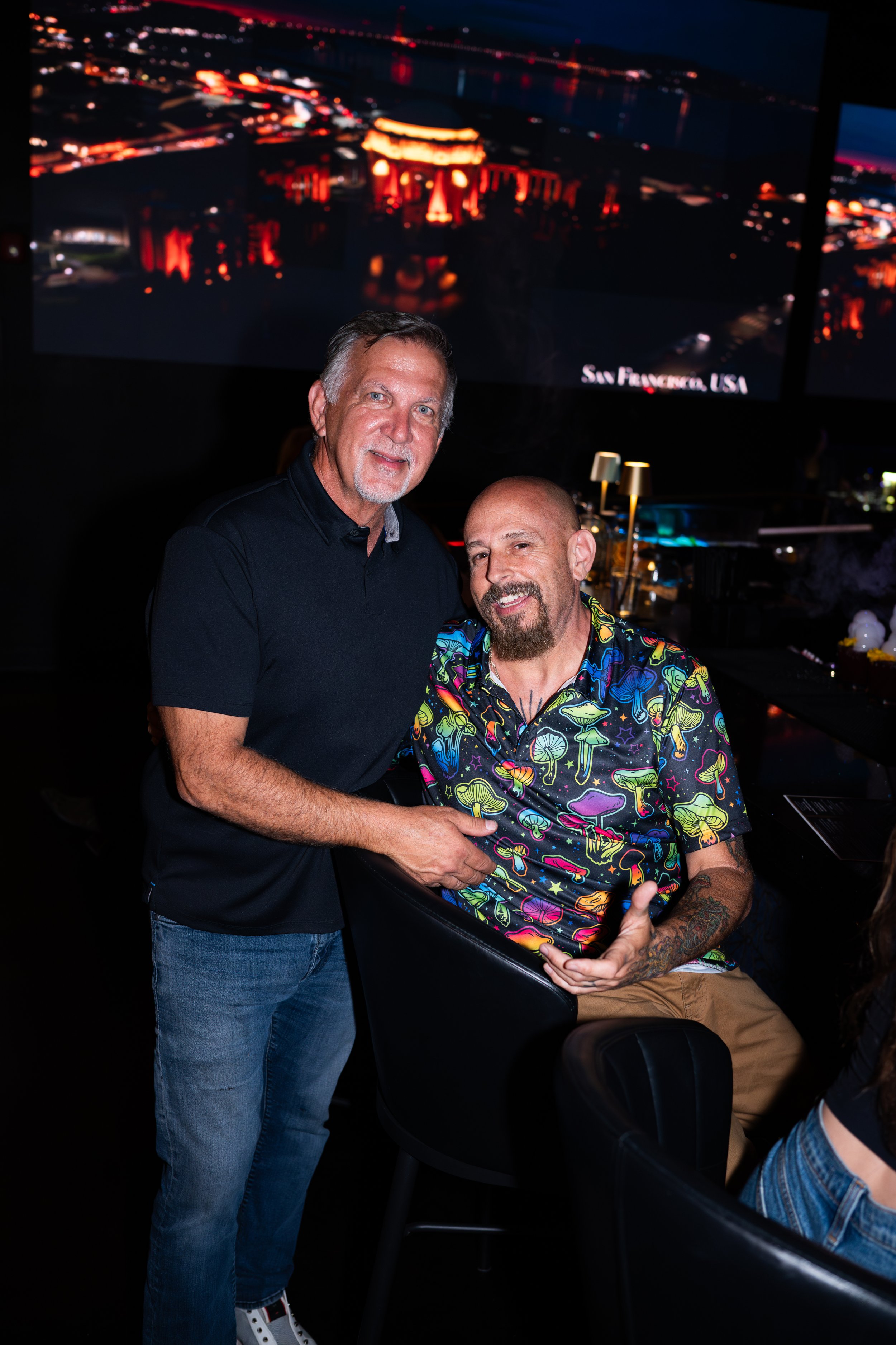 Two men shaking hands, one with gray hair and a beard wearing a black shirt, and the other bald with a goatee wearing a colorful mushroom-patterned shirt. They are in a dark indoor setting with large screens displaying a nighttime cityscape in the ba