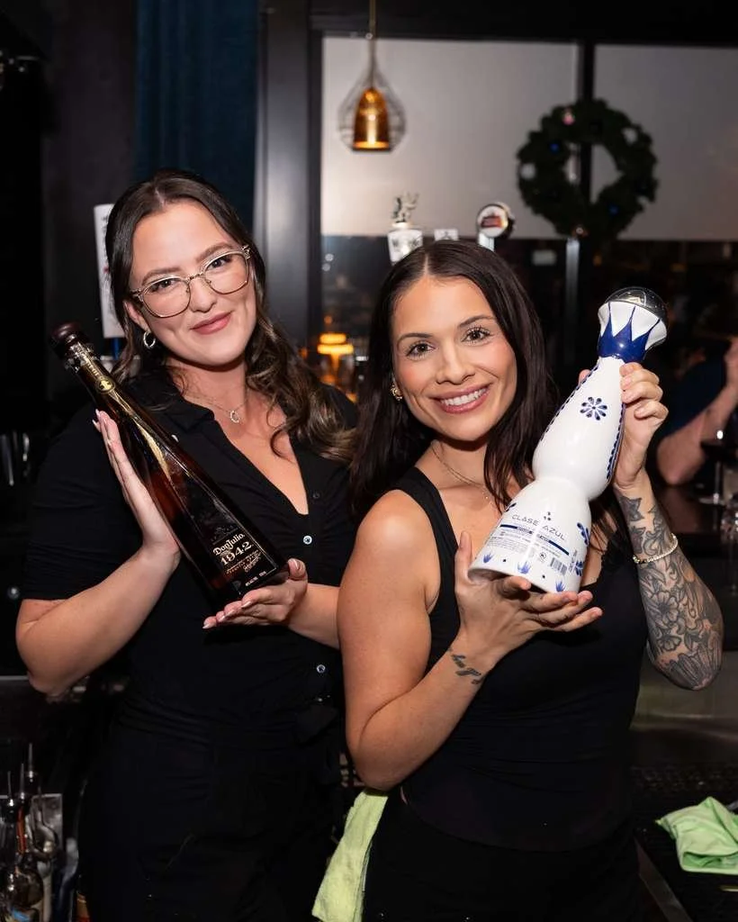 Two women smiling and holding decorative bottles inside a restaurant or bar with dark decor and a wreath in the background.