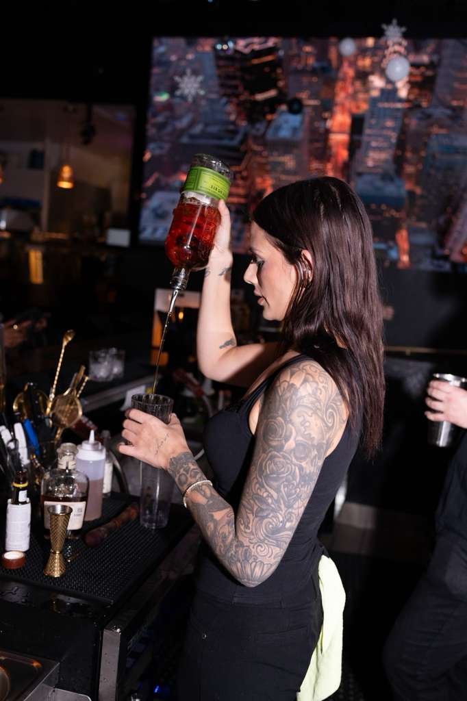 A woman with tattoos on her arm and shoulder, wearing a black tank top, pours a red drink from a large bottle into a glass at a bar. There is a cityscape with winter holiday decorations like snowflakes in the background.