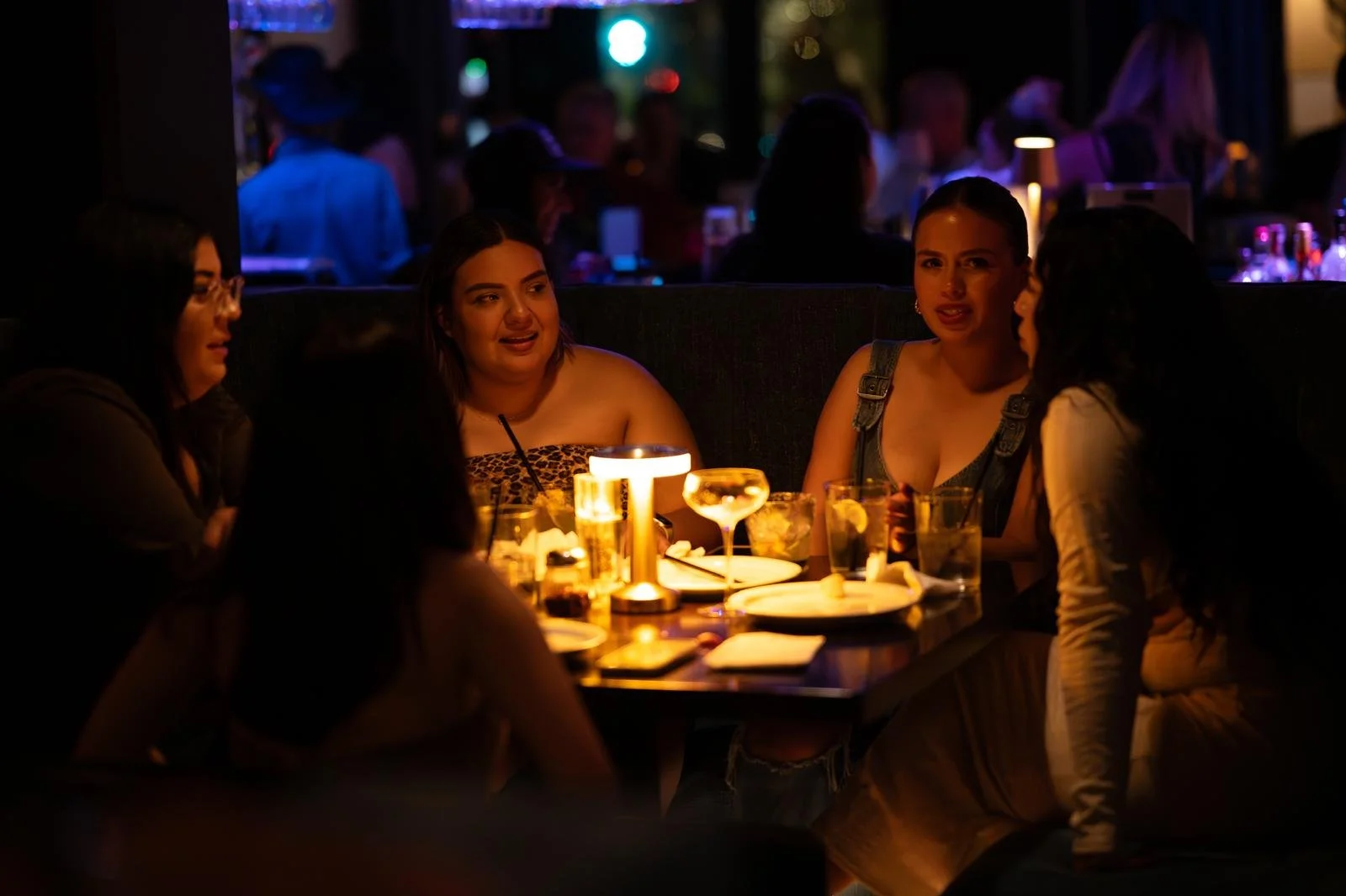 Four women sitting around a table at a restaurant or bar, engaging in conversation, illuminated by warm outdoor lighting with other people in the background.