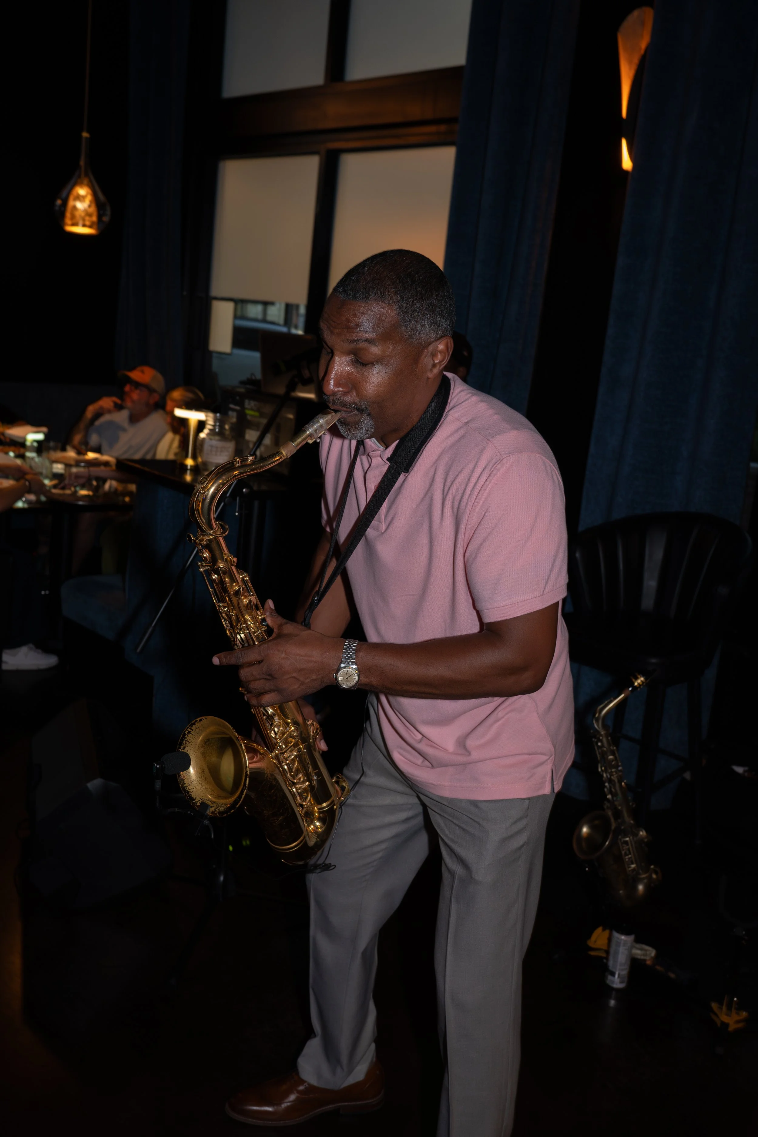 Man playing a gold saxophone indoors at a social gathering, with dark curtains and other people seated at tables in the background.