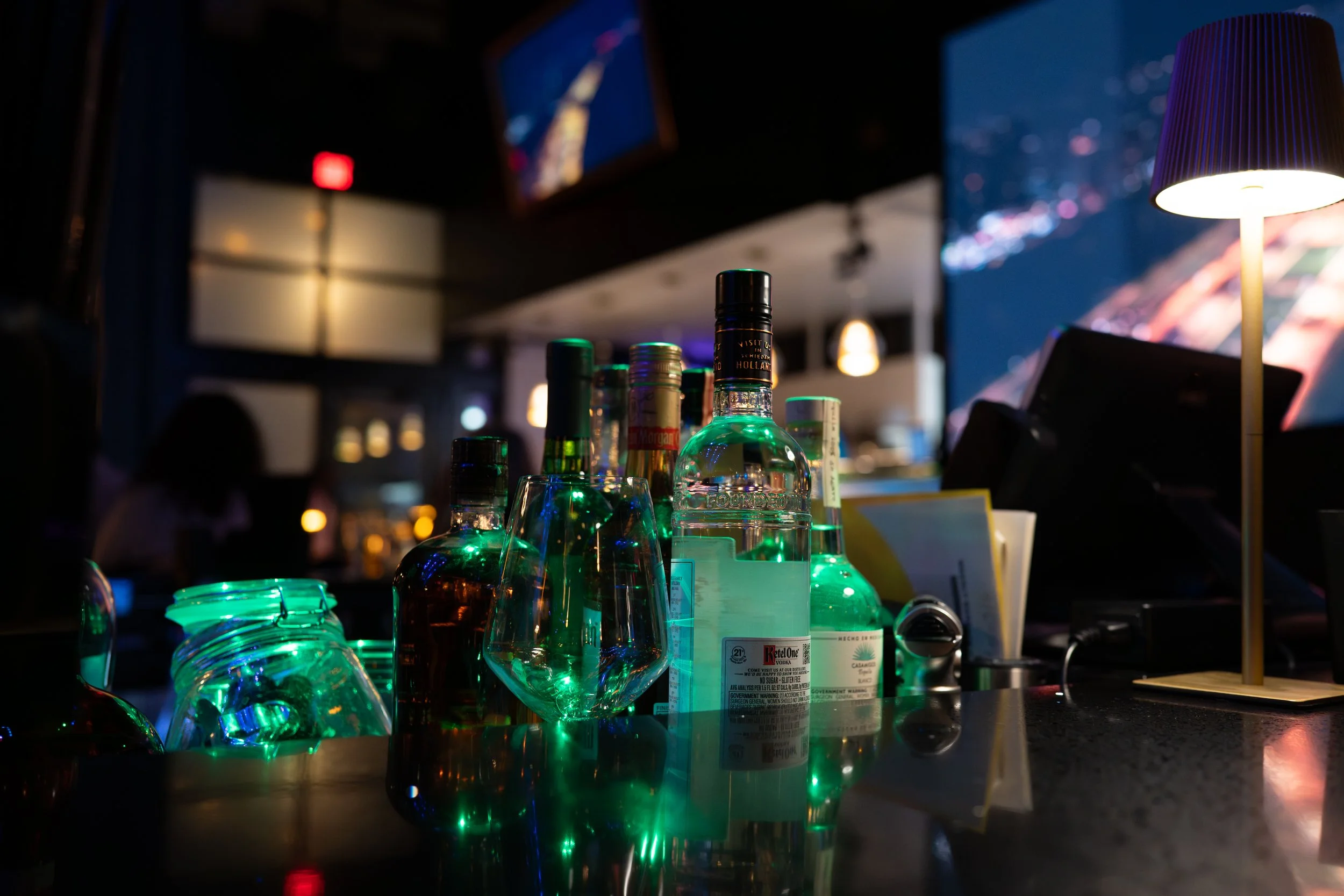 Several bottles of liquor on a bar counter illuminated with green light in a dimly lit bar or nightclub.