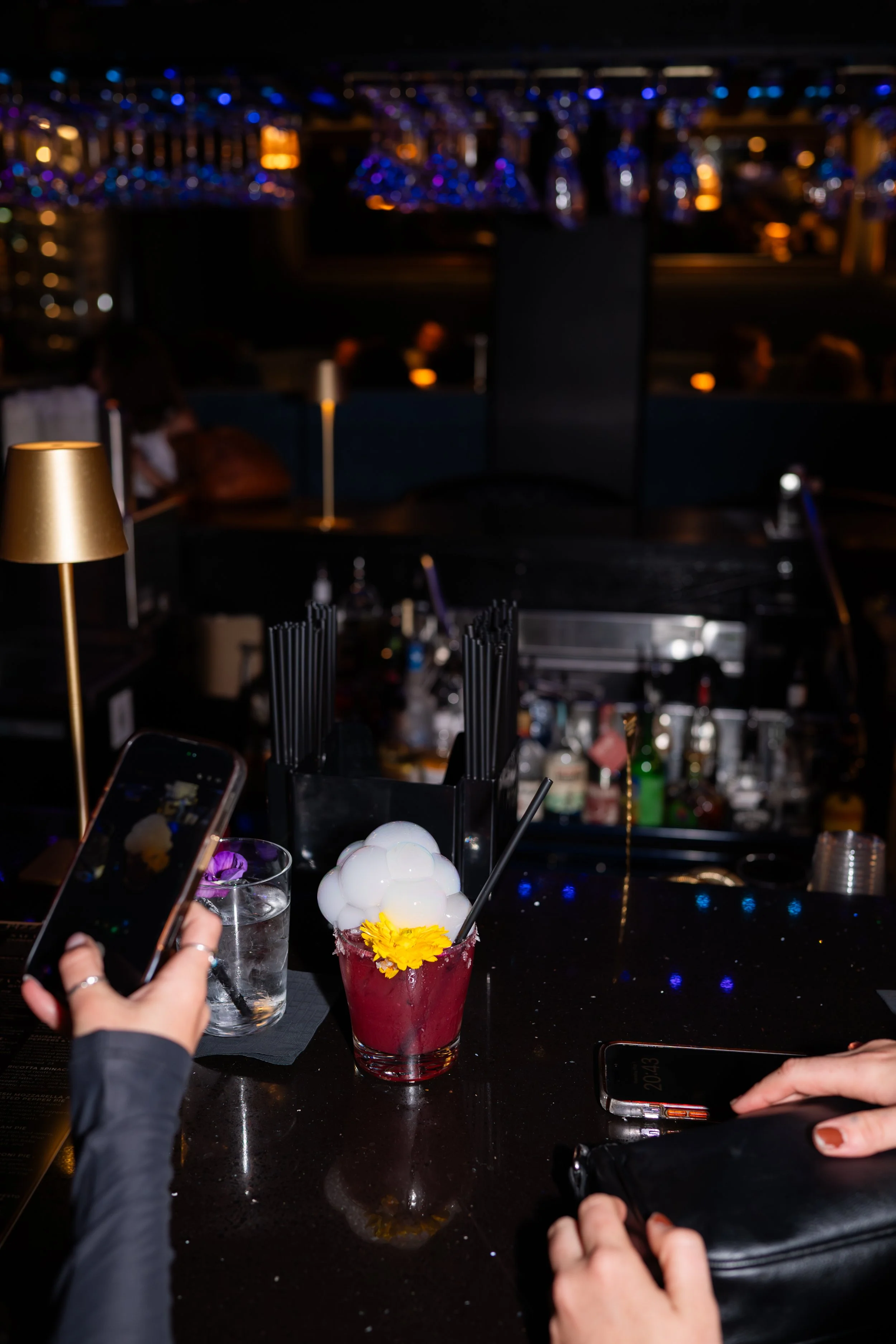 Close-up of a cocktail glass filled with a red drink garnished with a yellow flower, ice, and purple flower petals, on a black bar counter. Two people are visible, one taking a photo with a smartphone and the other holding a black purse. The backgrou