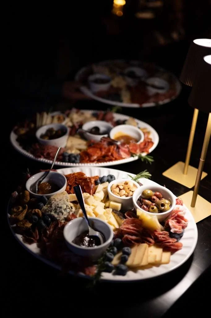 Charcuterie board with cheeses, olives, nuts, cured meats, grapes, and dipping sauces, illuminated by a gold candlestick lamp in a dark setting.