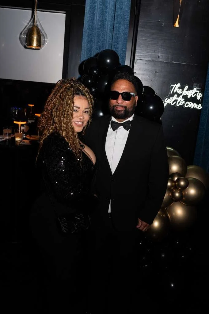 A woman and a man dressed in formal attire posing together at a celebration, with black and gold balloons and a neon sign in the background.