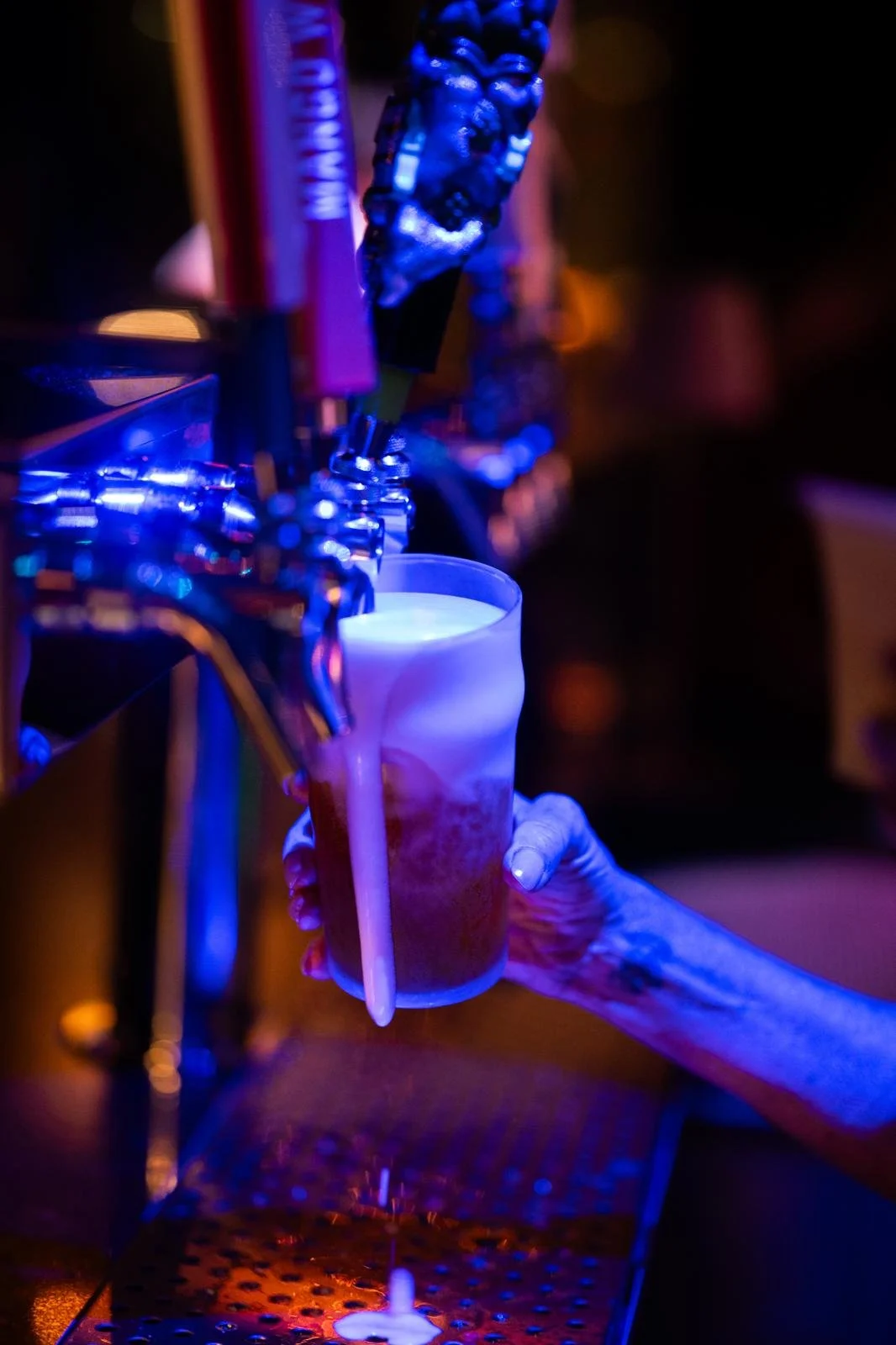 A hand holding a glass under beer taps at a bar counter with colorful blue and purple lighting.
