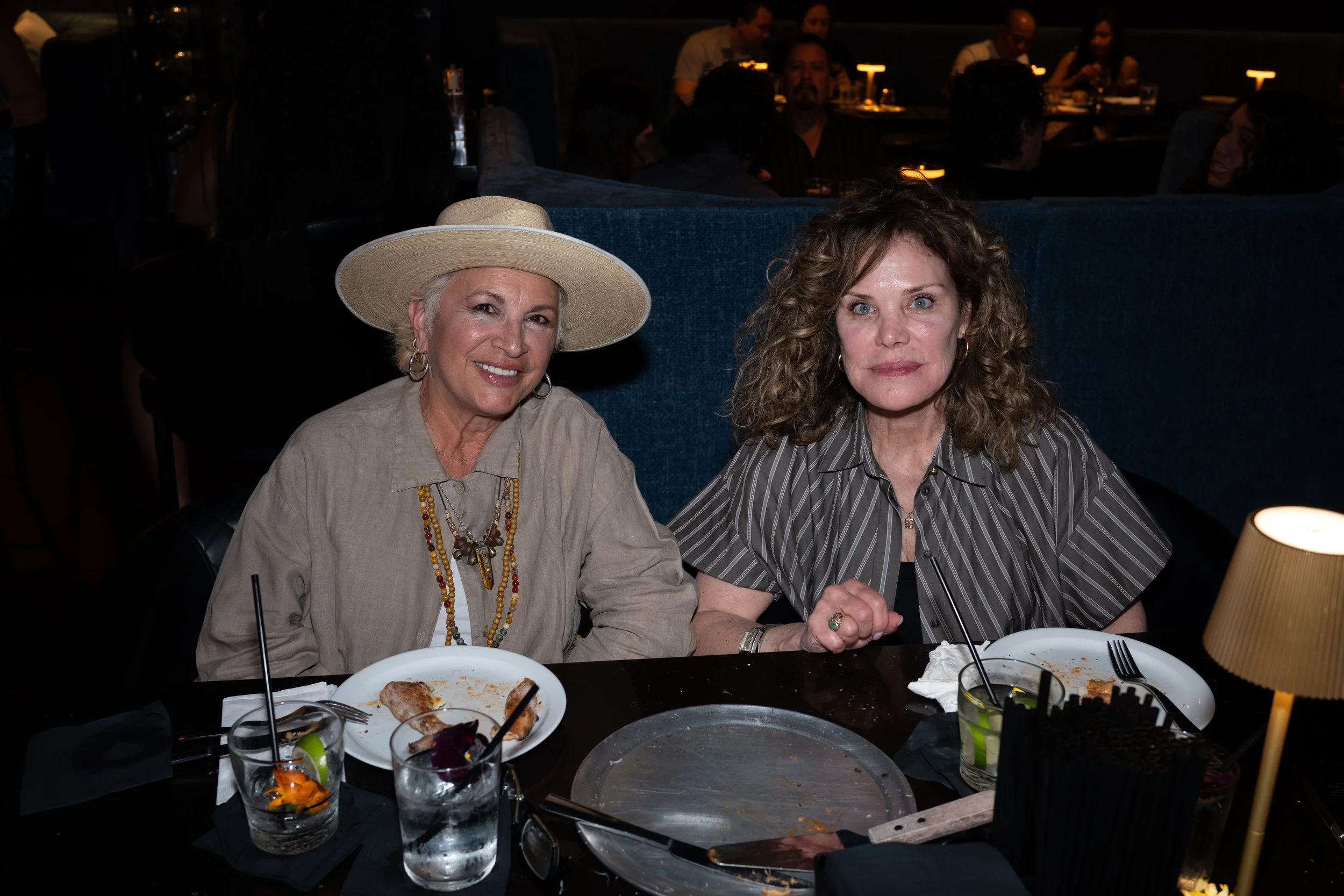 Two women sitting at a restaurant table, one wearing a large straw hat and jewelry, the other with curly hair and a striped shirt. There are partially eaten plates of food, glasses of drinks with lime slices, and tableware on the table.