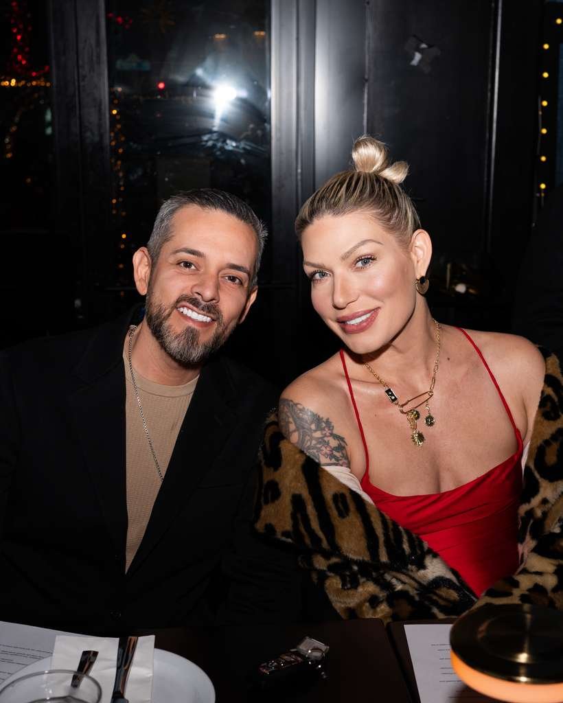A smiling man with a beard and a woman with a top bun, wearing a red dress and a leopard print shawl, sitting at a table in a dimly lit restaurant or bar at night.