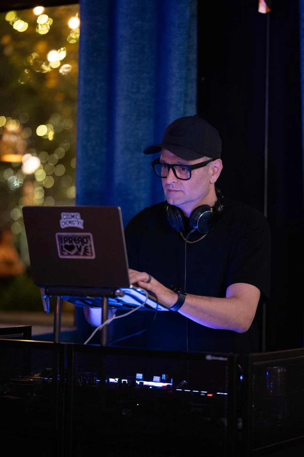 A DJ wearing a black cap, glasses, black t-shirt, and headphones around his neck, working with a laptop in a dimly lit setting with colorful bokeh lights in the background.