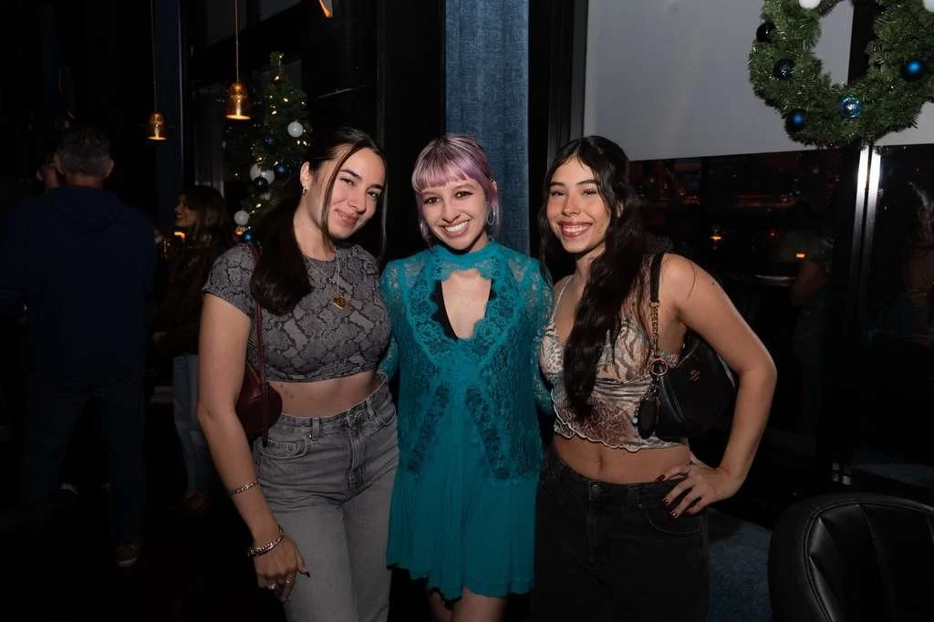 Three women smiling at a festive event with Christmas decorations in the background, including a Christmas tree with ornaments and hanging lights.
