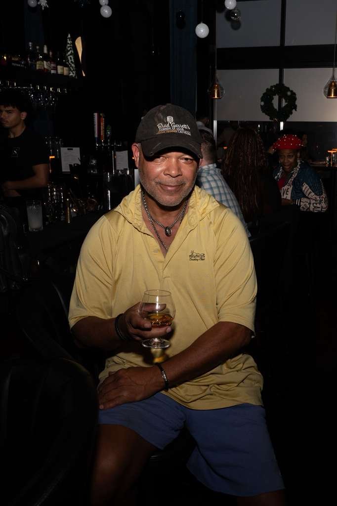 A man sitting at a bar holding a glass of whiskey, wearing a yellow polo shirt, a black baseball cap, and a necklace, with a dimly lit bar and other patrons in the background.