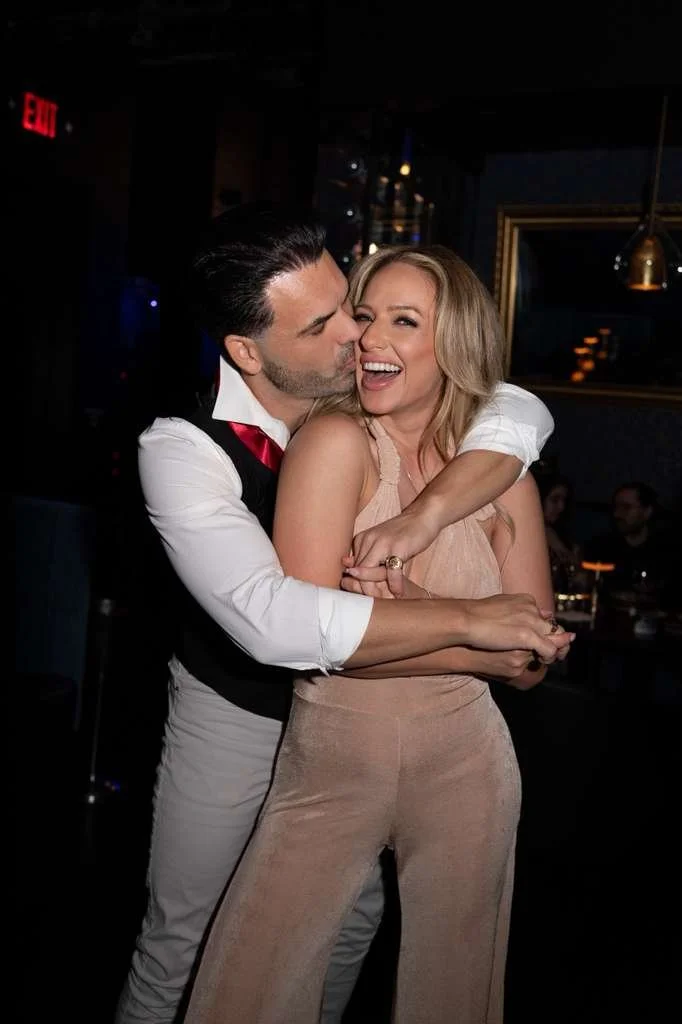 A man and woman are happily hugging and smiling in a dimly lit indoor setting, possibly a bar or restaurant.