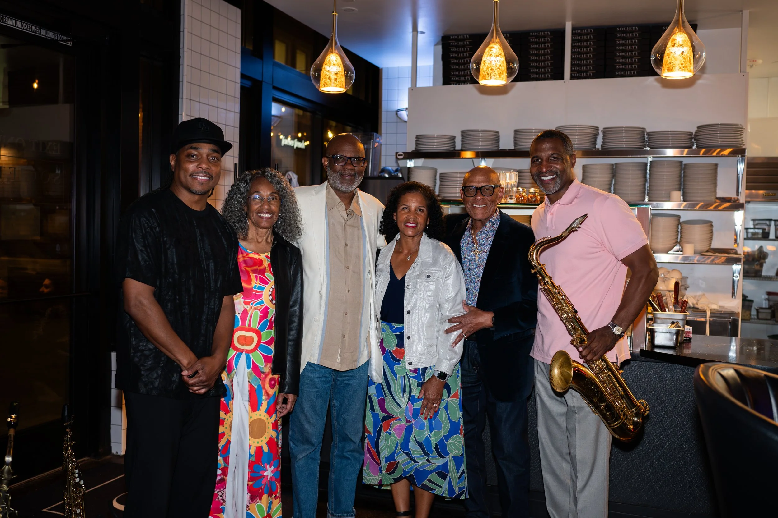Group of six diverse people smiling in a restaurant kitchen, with shelves of plates in the background. One man is holding a saxophone.