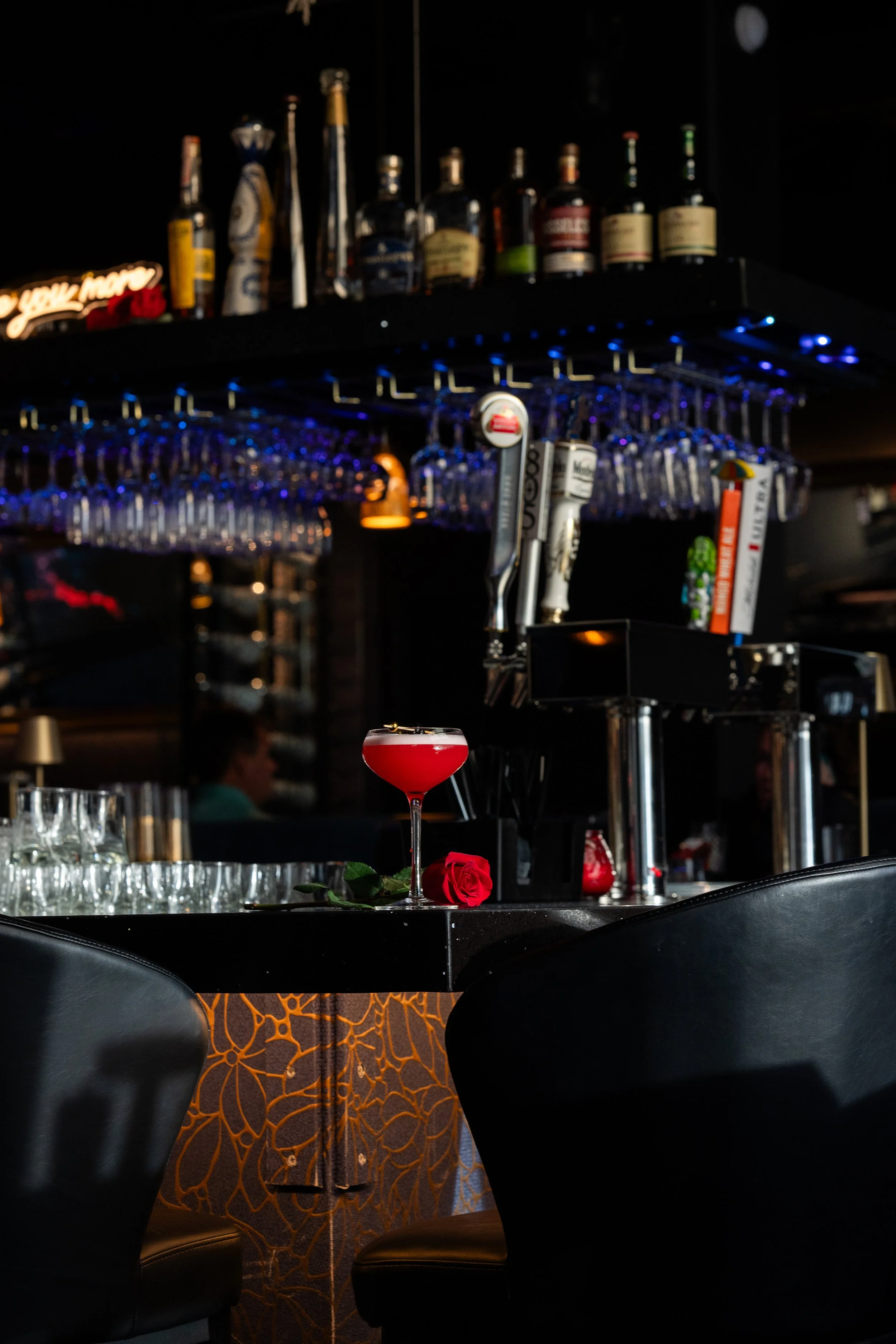 A dark bar with a red cocktail and a red rose on the counter, illuminated by soft lighting, with hanging glasses and liquor bottles in the background.