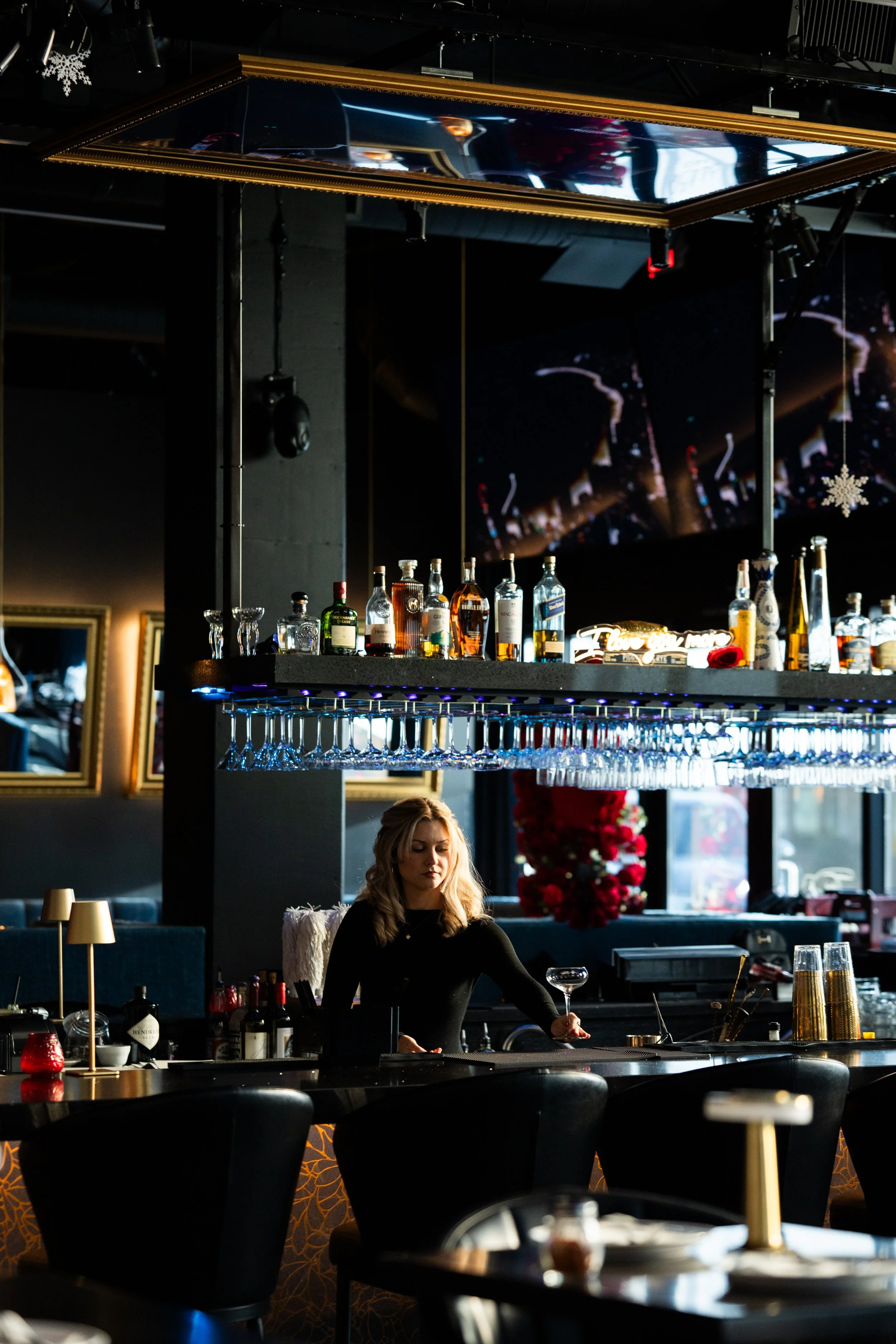 A woman behind a bar counter in a dimly lit restaurant or bar, with bottles of alcohol and glassware displayed above her. There are decorative elements like mirrors and framed pictures on the dark-colored walls.