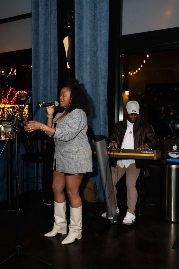 A woman singing into a microphone with a man playing a keyboard behind her at a dimly lit indoor venue, with blue curtains and city lights visible through the windows.