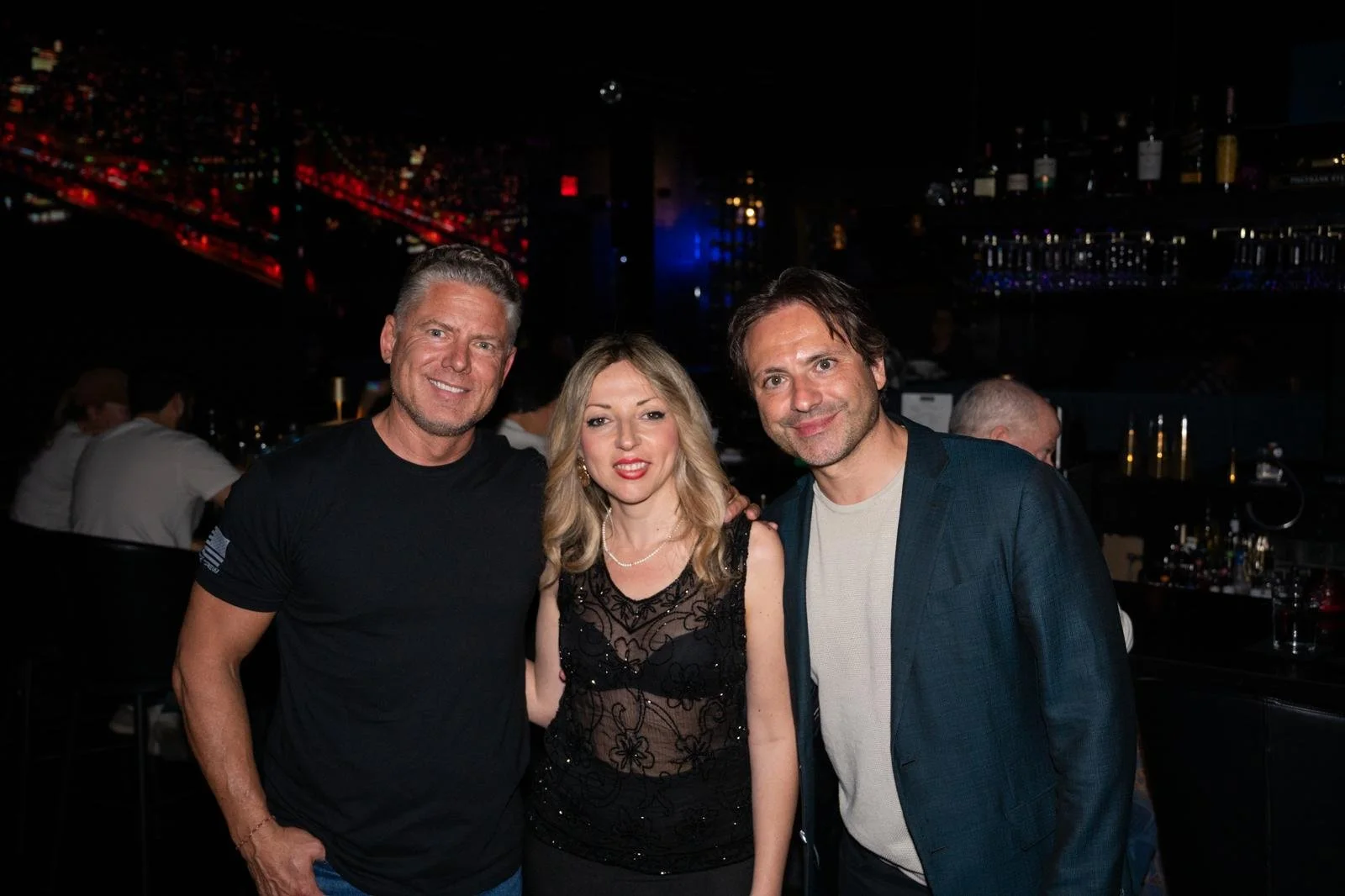 Three people smiling at a social event in a dimly lit bar, with a cityscape backdrop visible in the background.
