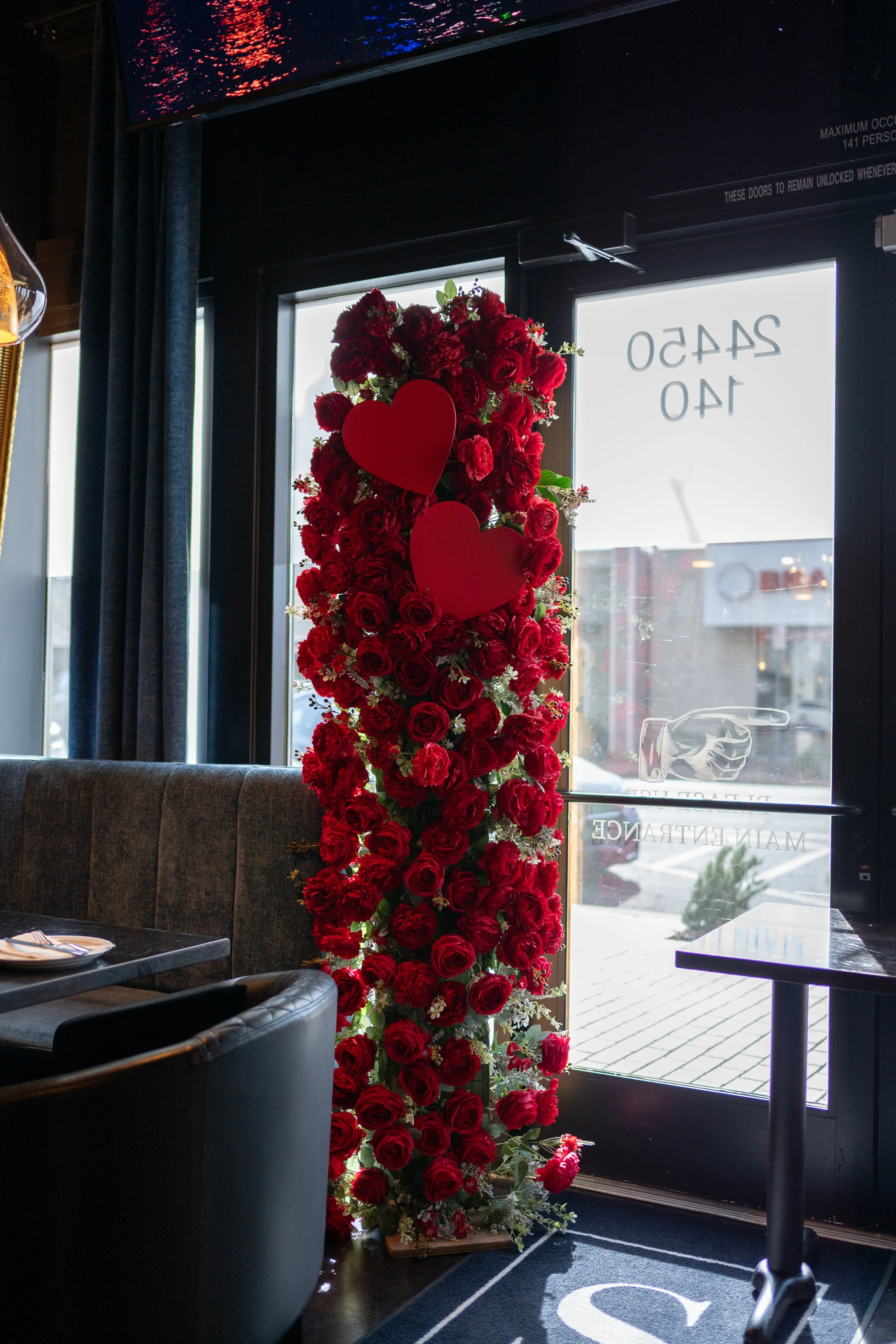A tall standing display of red roses with two large red heart cutouts, positioned near a glass door inside a restaurant or cafe.