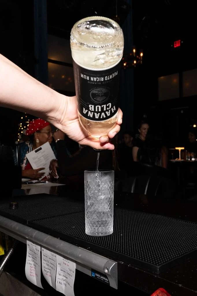 Person pouring a bottle of Haassana Club sparkling wine into a glass at a bar, with people in the background.