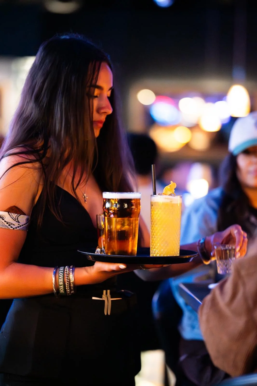 A woman with long dark hair holding a black tray with a glass of beer and a yellow cocktail at a nighttime outdoor bar or restaurant.