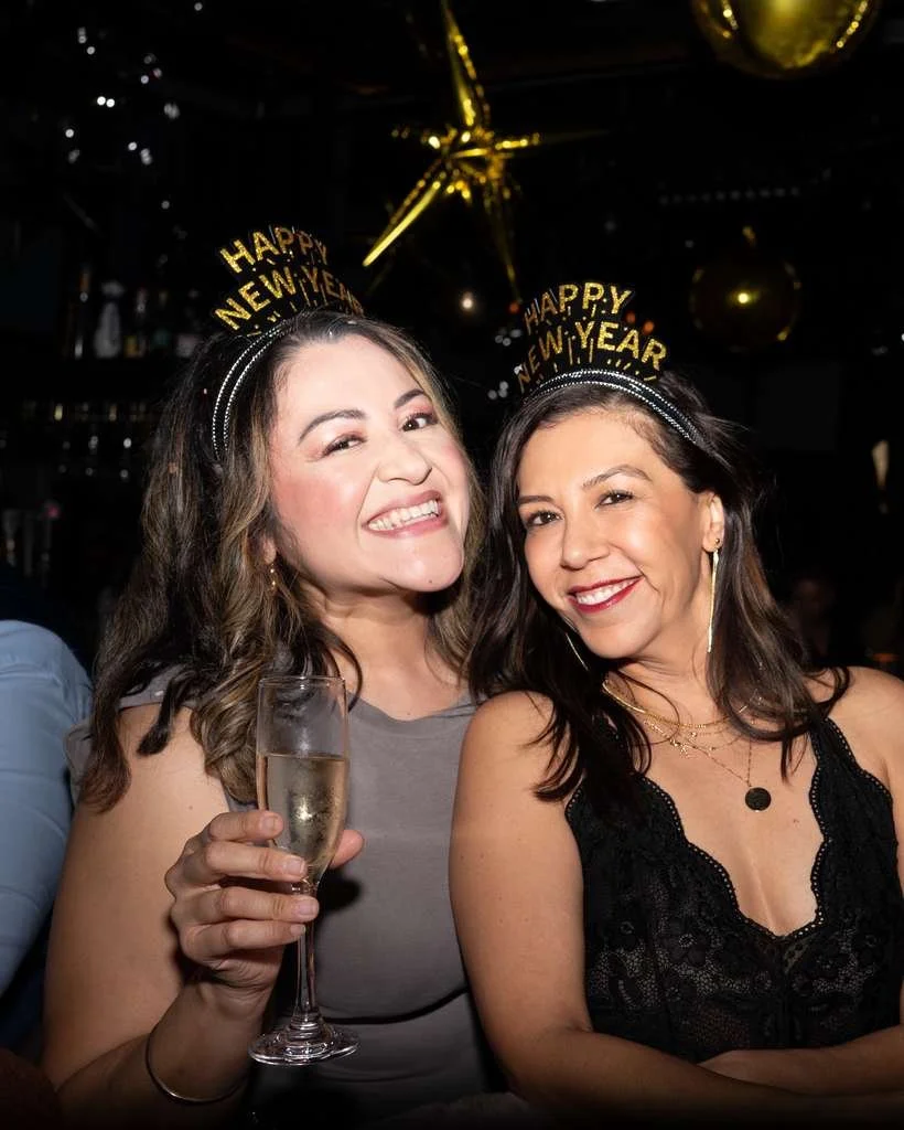 Two women smiling at a New Year's celebration wearing 'Happy New Year' headbands, one holding a glass of champagne, in a festive setting with black background and gold decorations.