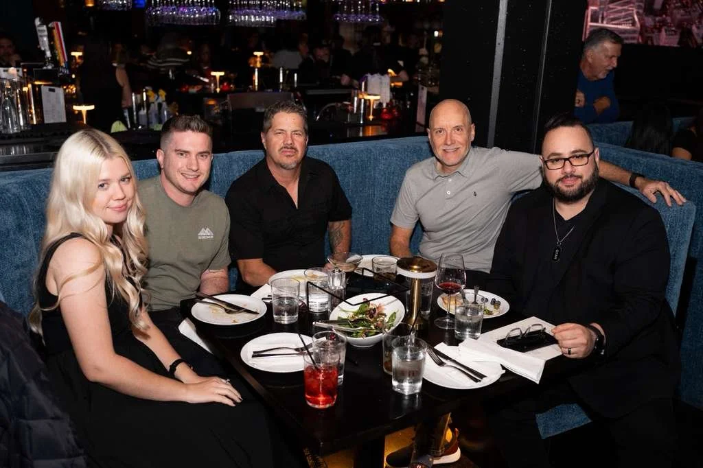 Group of five people sitting at a restaurant table with plates, glasses, and a salad, in a dimly lit dining area.