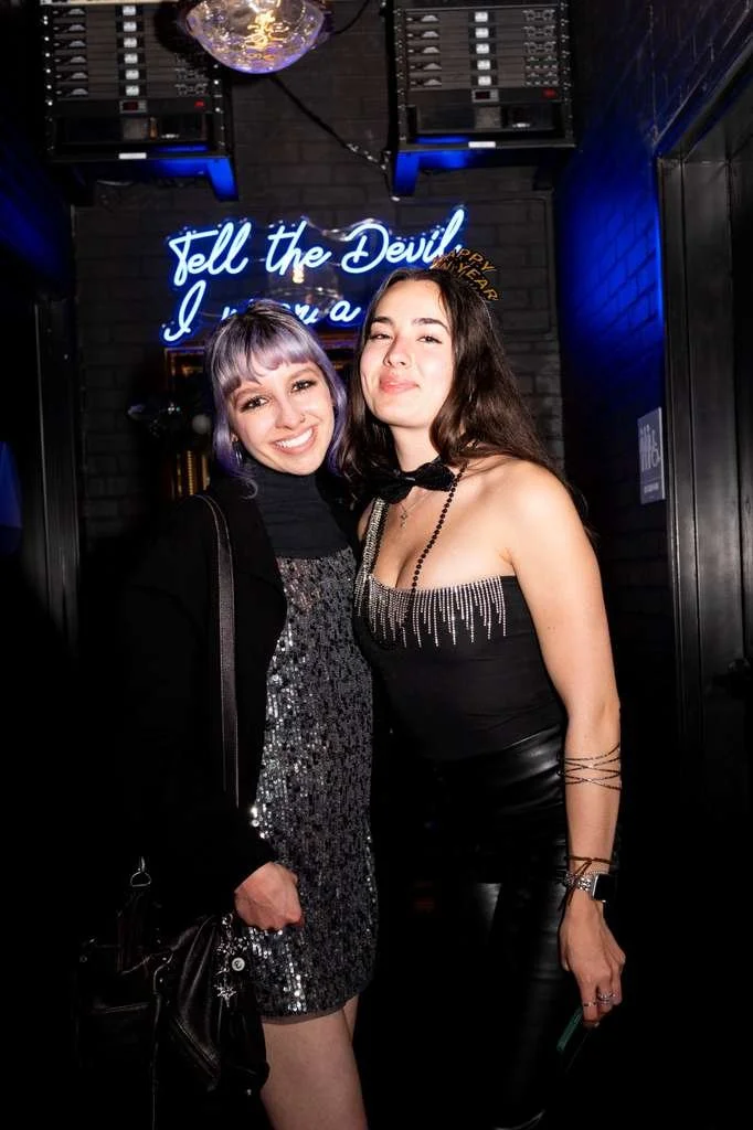 Two women smiling and posing for a photo in a nightclub or bar with a neon sign behind them that reads "Tell the Devil I".