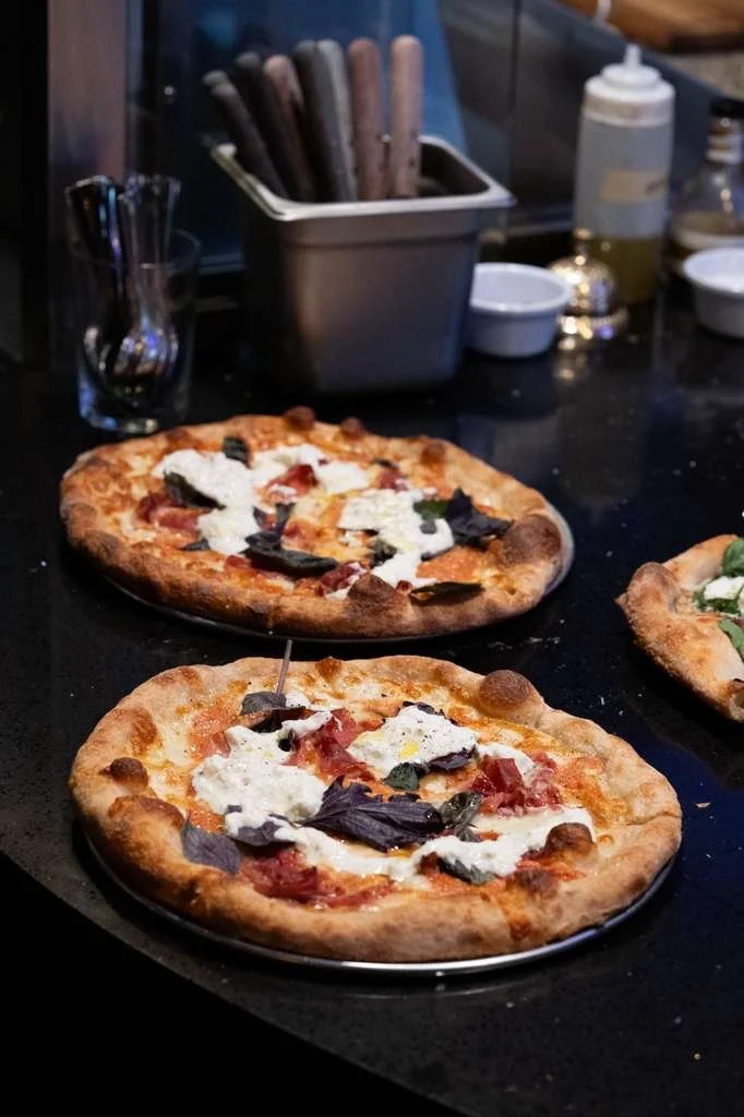 Two personal pizzas with cheese, tomato, basil, and dark leafy toppings on a black surface in a pizzeria kitchen.