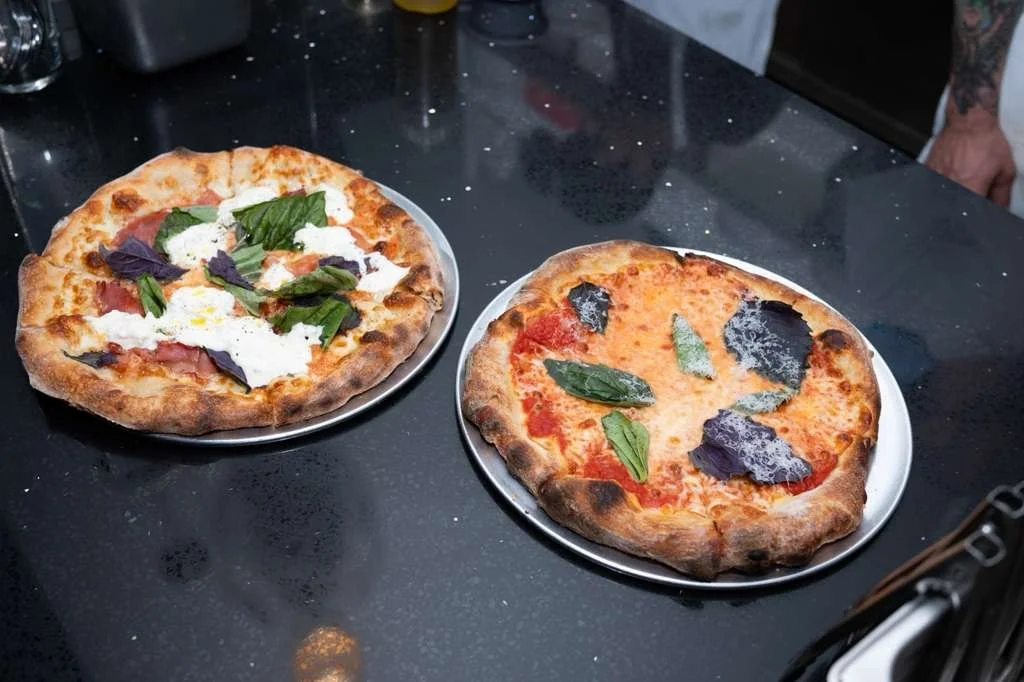 Two pizzas on metal plates on a black counter with some scattered white flour or salt. The pizza on the left has a crust with cheese, ham, fresh basil, and ricotta cheese. The pizza on the right has melted cheese with basil and purple basil leaves.