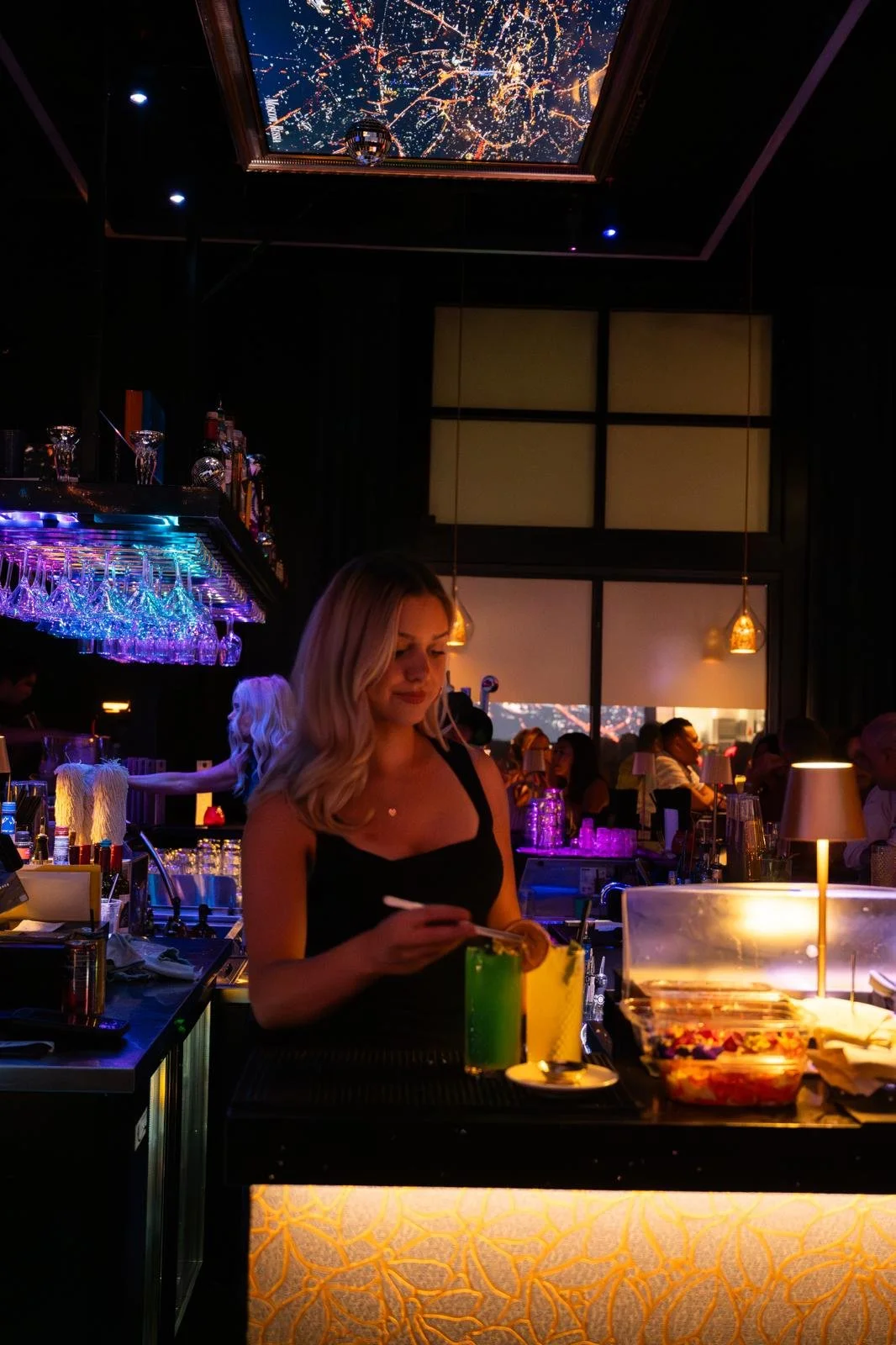 A woman behind a bar counter in a dimly lit, vibrant nightclub with colorful mood lighting, barware, and people in the background.