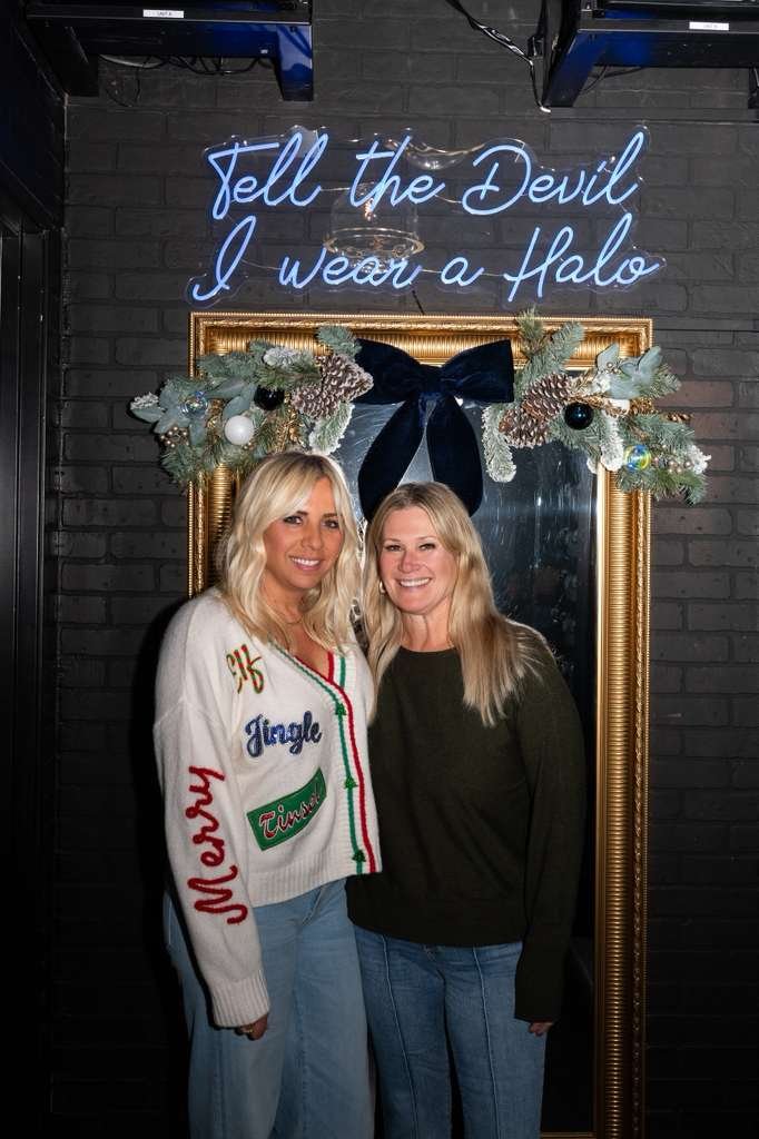 Two women standing in front of a mirror with a gold frame and Christmas decorations, including pinecones and ornaments. A neon sign above reads, "Tell the Devil I Wear a Halo." One woman is wearing a Christmas sweater with festive words and designs, 