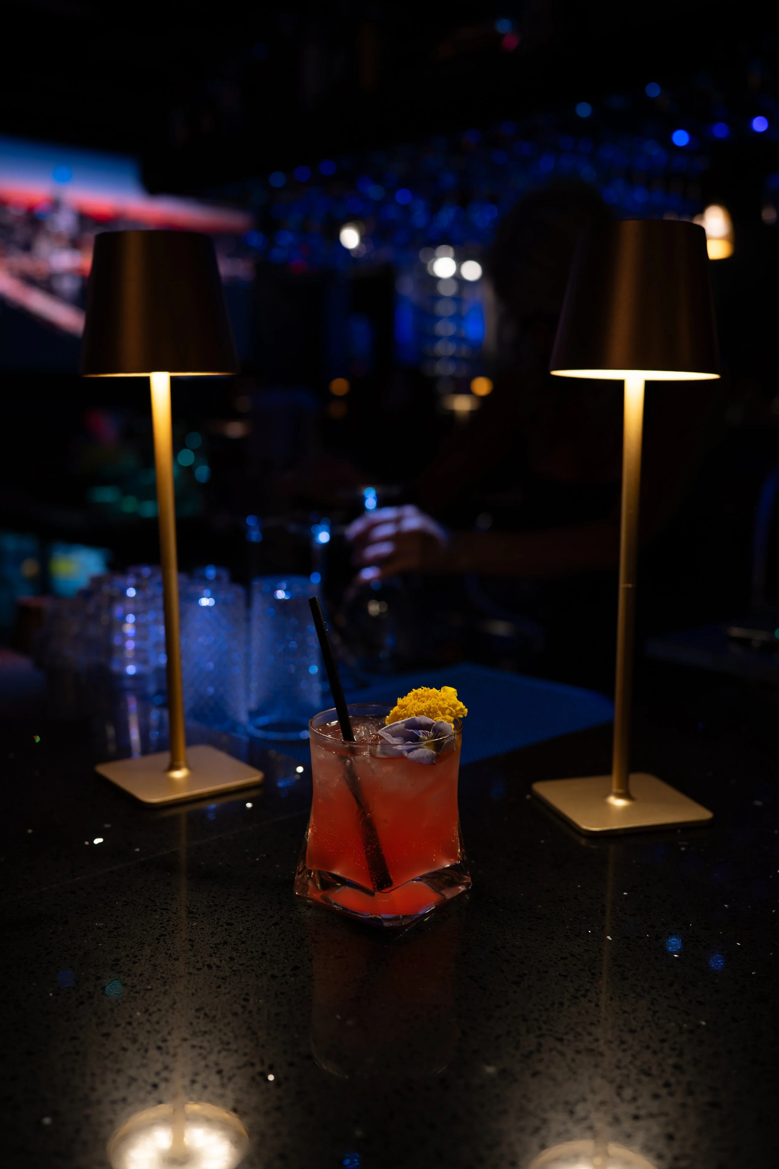 A dimly lit bar scene with two small table lamps casting warm light on a black countertop. In the foreground, a pink cocktail garnished with flowers and a lemon is visible. The background features blurred blue and white lights, a person holding a gla