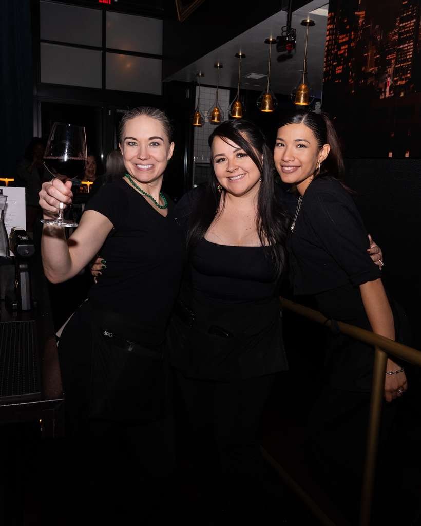 Three women smiling and posing together inside a modern restaurant or bar, with one holding a glass of red wine, illuminated by warm pendant lighting.