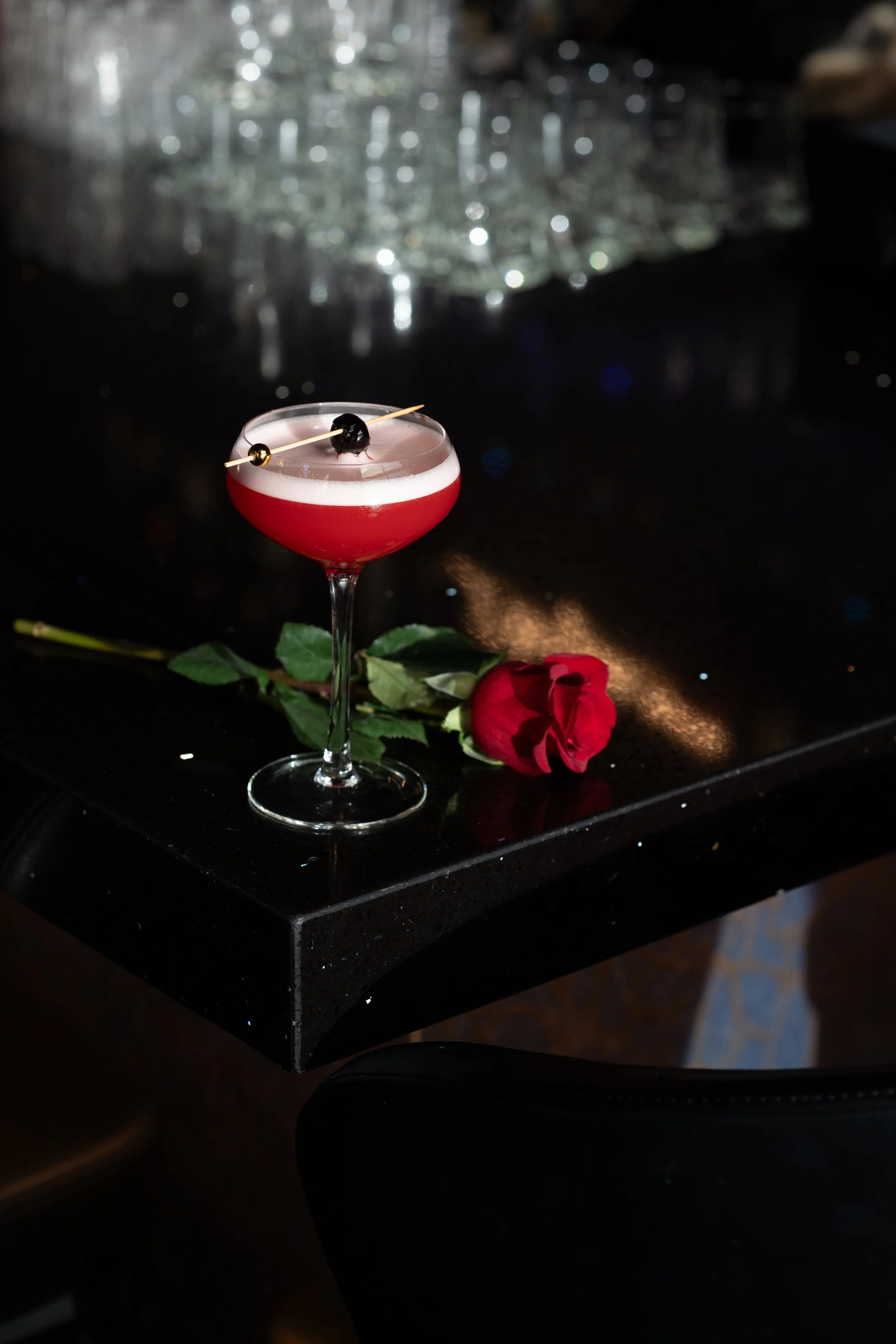 A pink cocktail with a cherry garnish and two small gold balls on a toothpick, resting on a black table with a red rose beside it, in a dimly lit setting.
