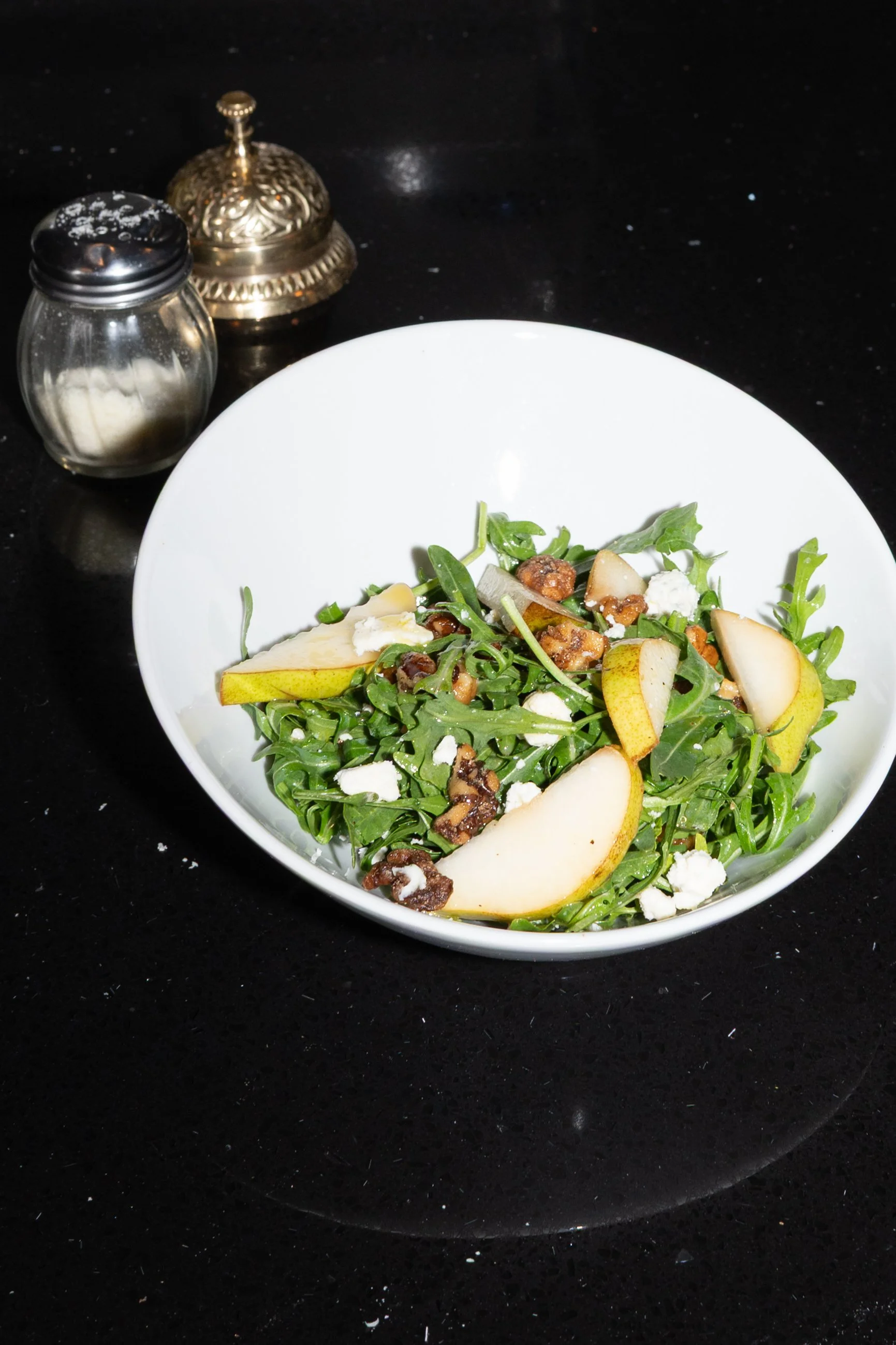 A salad with arugula, pear slices, walnut pieces, and crumbled cheese in a white bowl on a black surface.